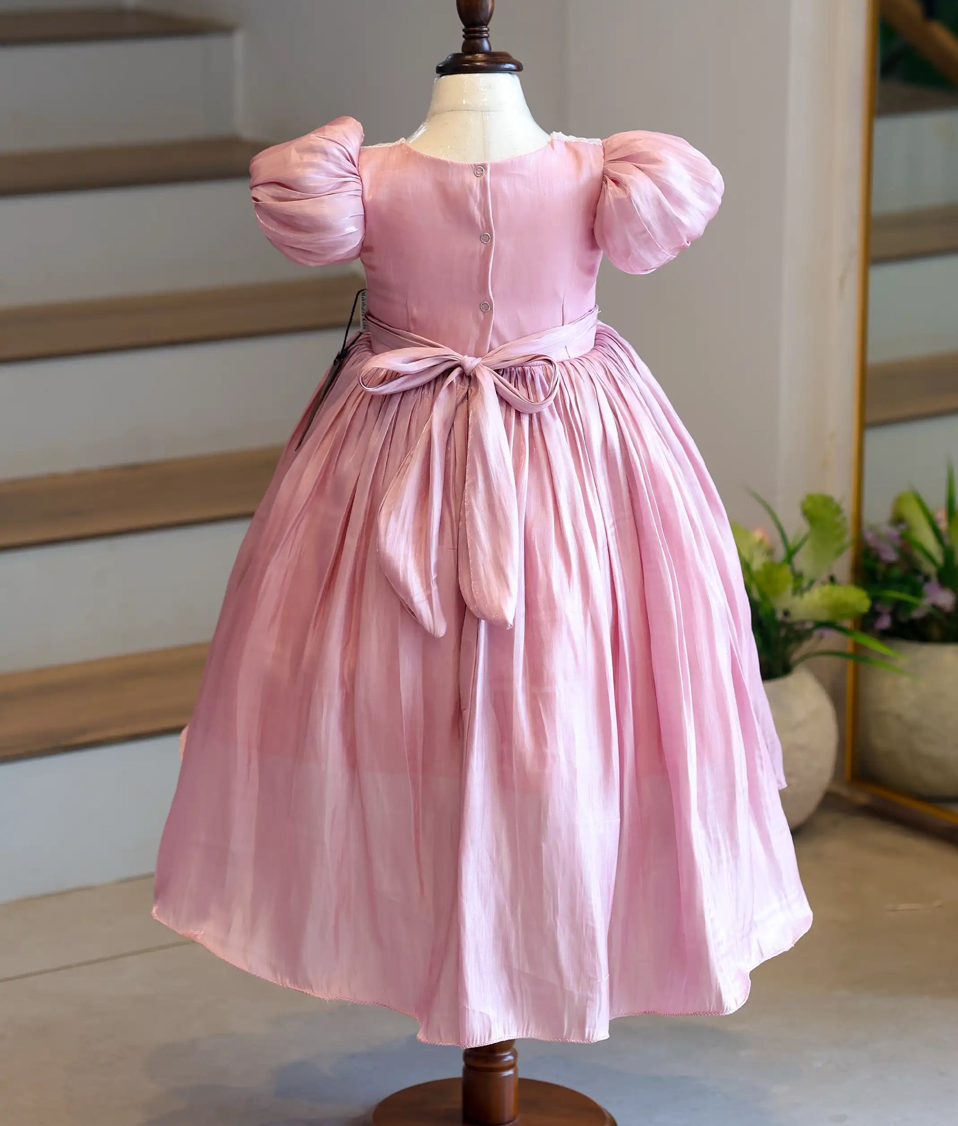 Back view of the kids' onion-pink party frock on a mannequin, displaying the symmetrical button closure and the neatly tied matching fabric sash bow at the waist.