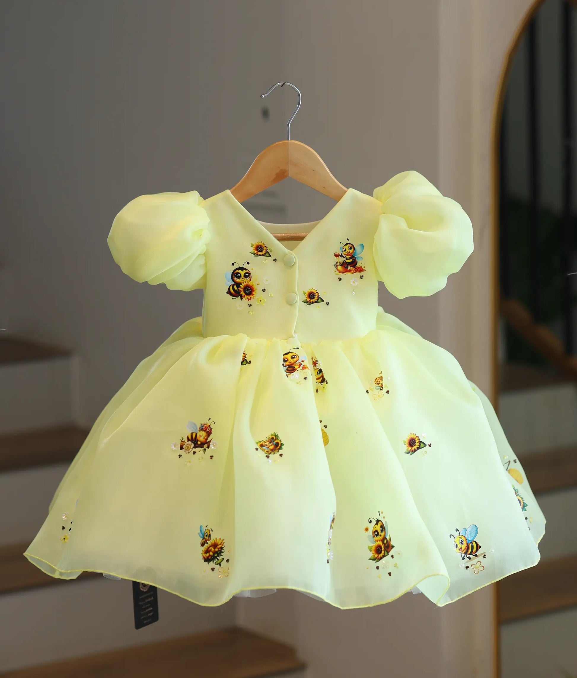 Back view of the honeybee-themed kids' dress on a hanger, displaying the V-cut back neckline, neat button closures, and printed bee motifs on the shoulders.
