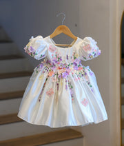 Full front view of a premium off-white satin kids' party frock hanging on a wooden hanger, covered in dense 3D pastel flowers and embroidered butterflies.