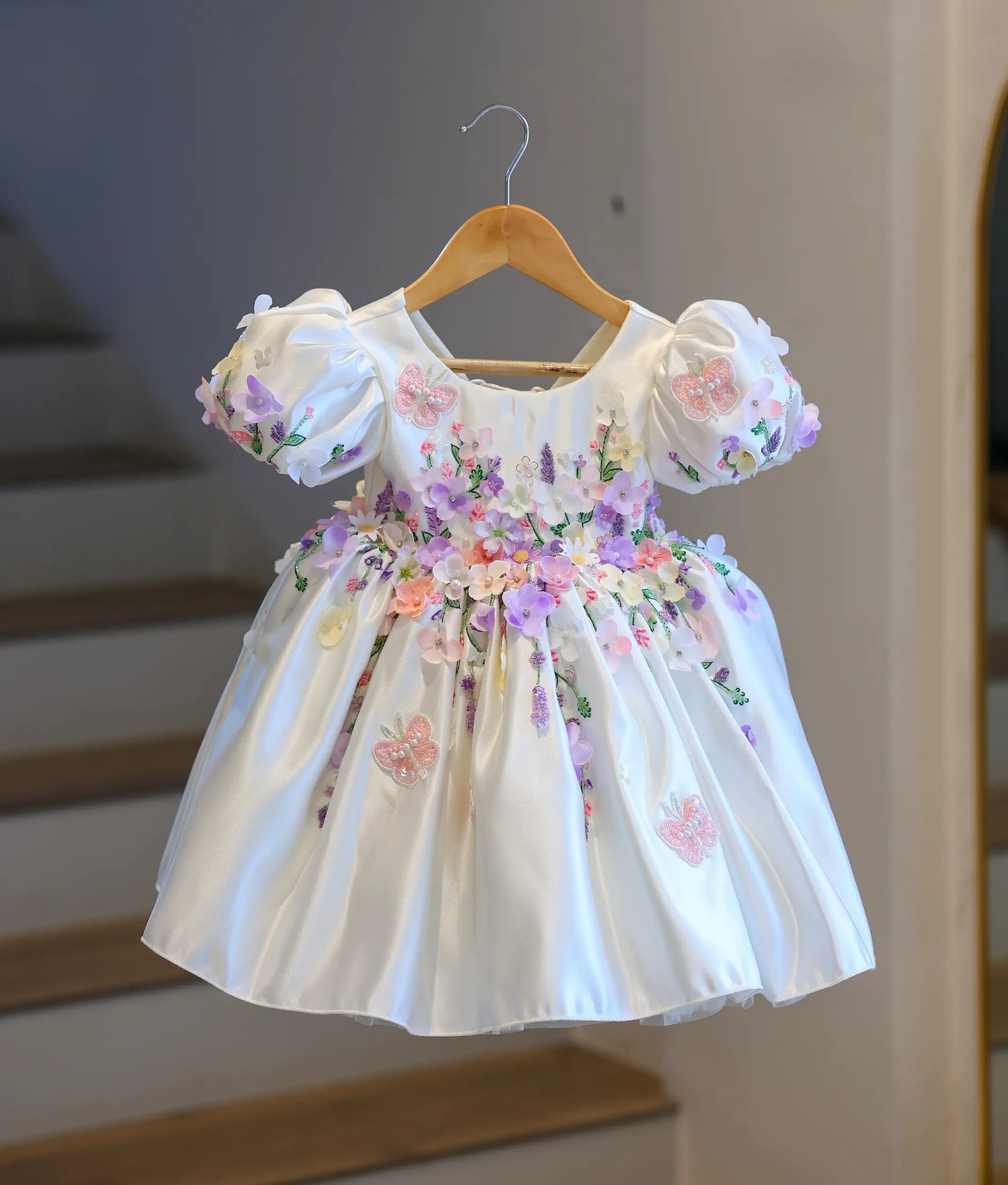 Full front view of a premium off-white satin kids' party frock hanging on a wooden hanger, covered in dense 3D pastel flowers and embroidered butterflies.