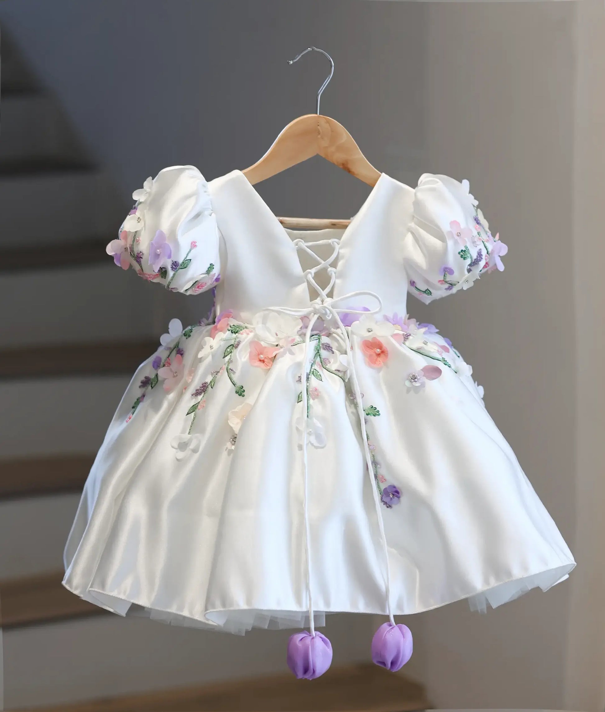 Full back view of the off-white kid's dress, displaying the elegant deep V-cut back, adjustable criss-cross corset satin strings, and hanging purple tulips.