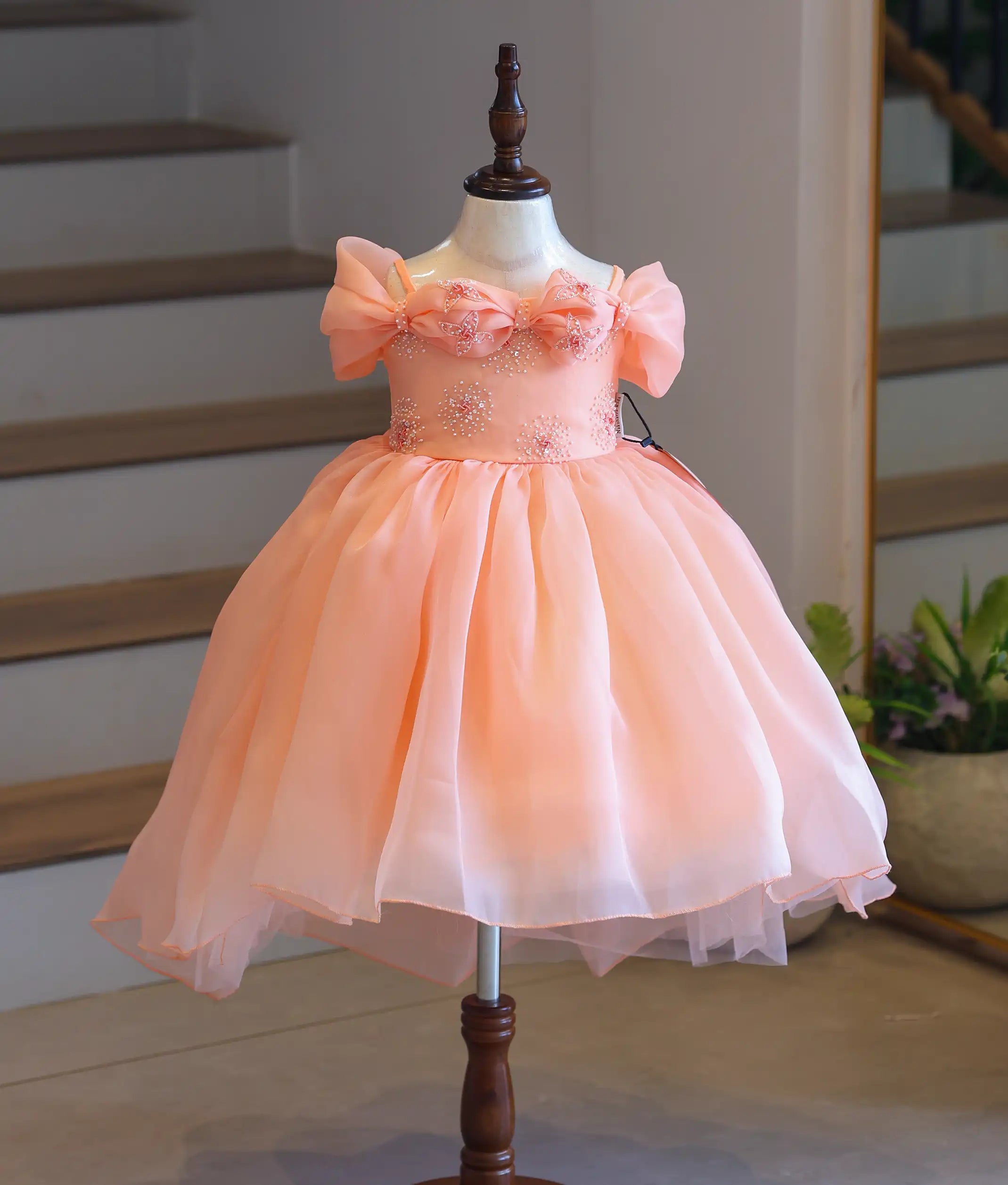Front view of a premium kids' peach organza party frock on a mannequin, showcasing the large statement bow on the top and flared skirt.