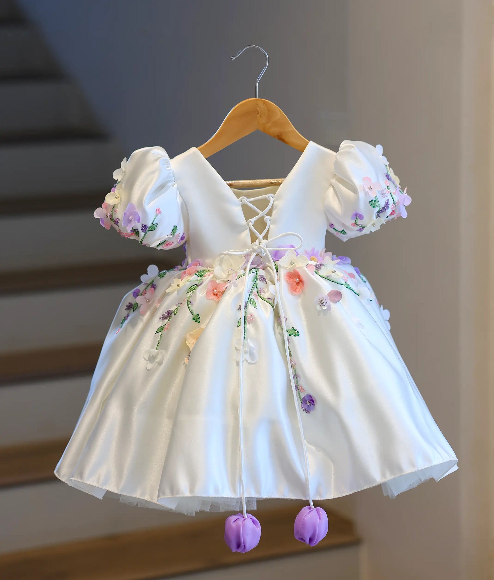Full back view of the off-white kids' dress, displaying the elegant deep V-cut back, adjustable criss-cross corset satin strings, and hanging purple tulips.