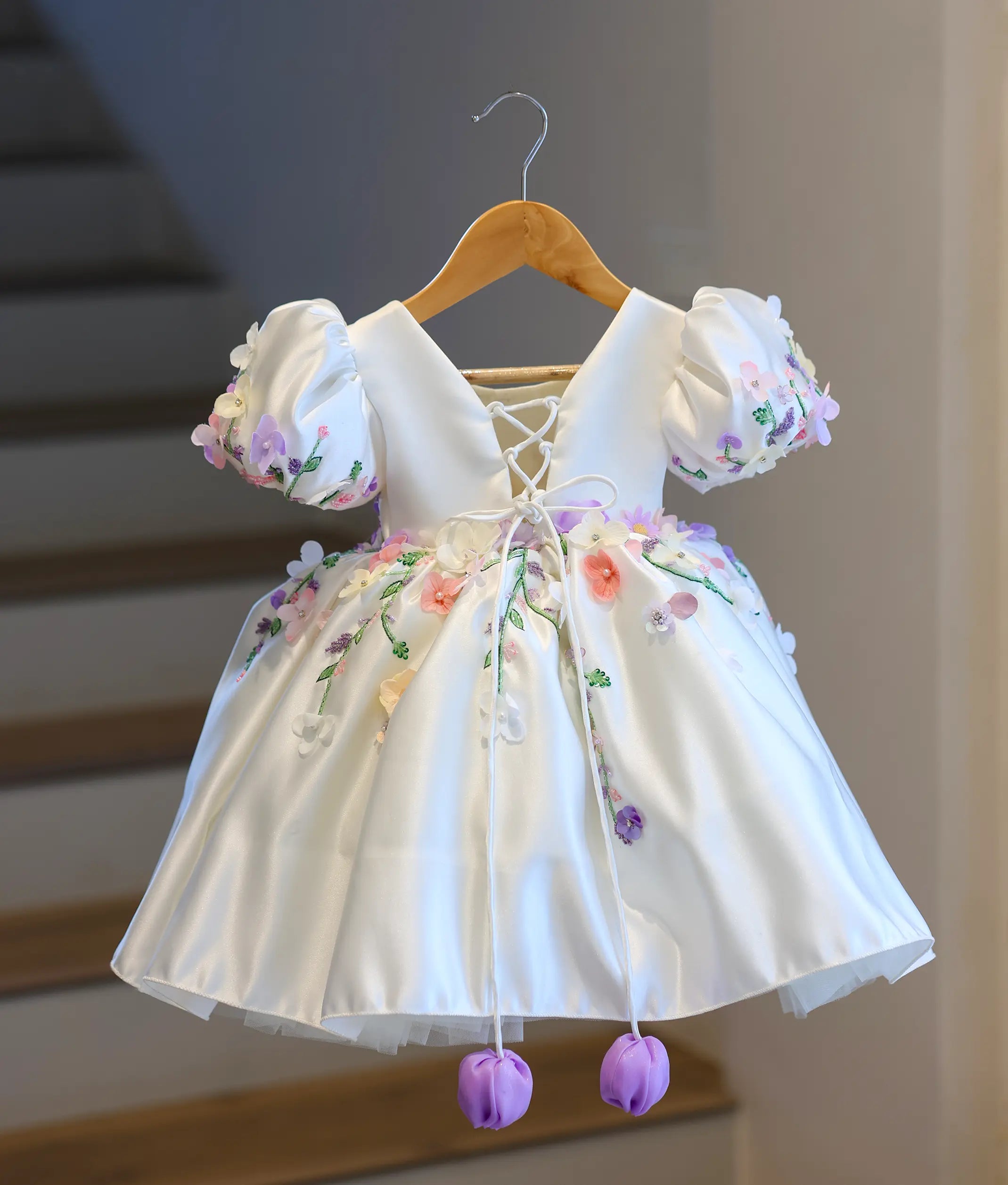 Full back view of the off-white kids' dress, displaying the elegant deep V-cut back, adjustable criss-cross corset satin strings, and hanging purple tulips.