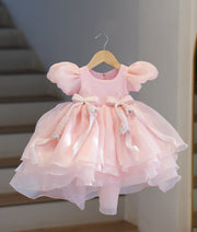 Front full view of baby pink shimmer organza birthday frock with a layered top, puffy sleeves, and flared tiered bottom.