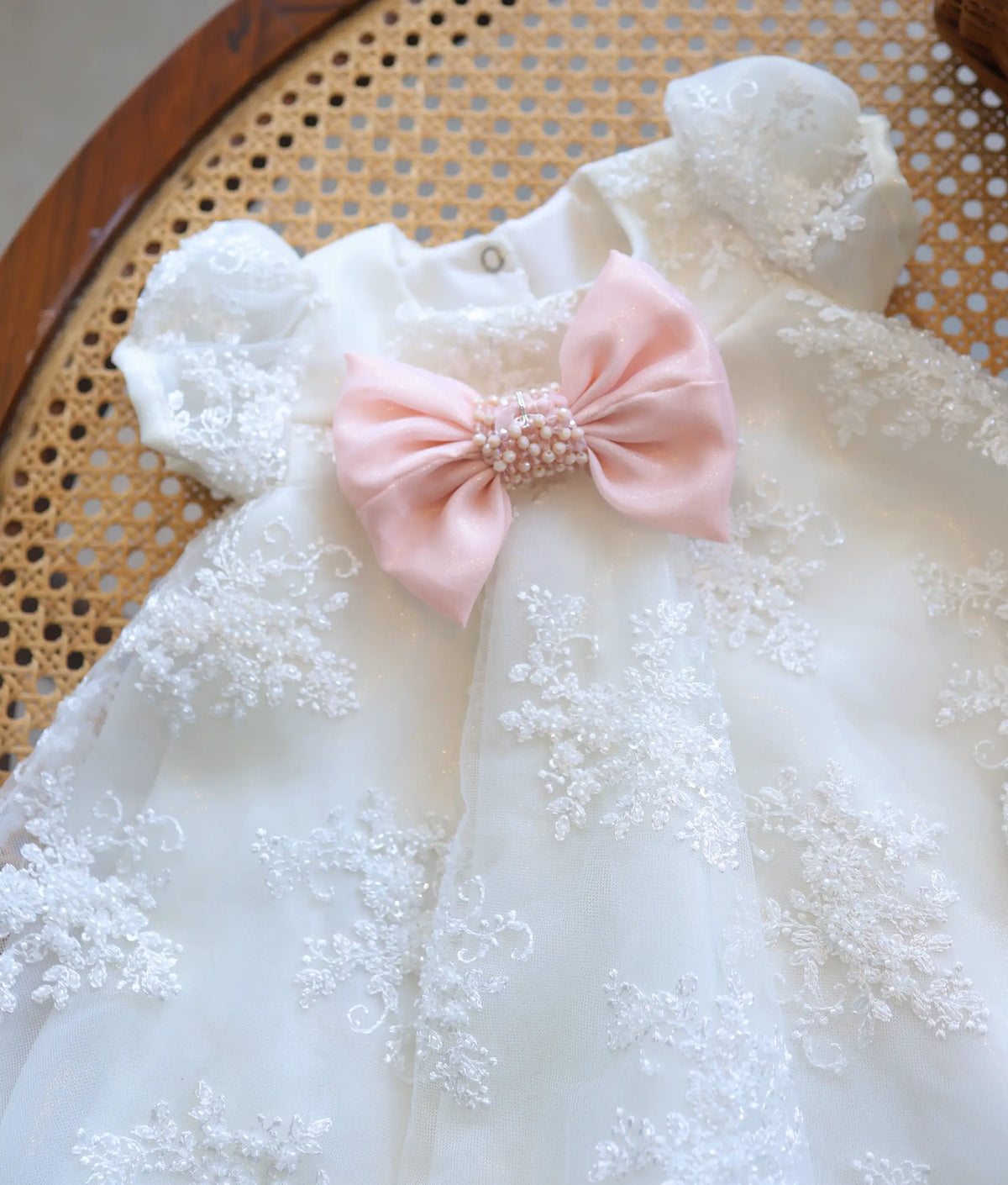Close-up of off-white baby girl baptism frock featuring delicate net embroidery and a large pastel pink bow with pearl center.