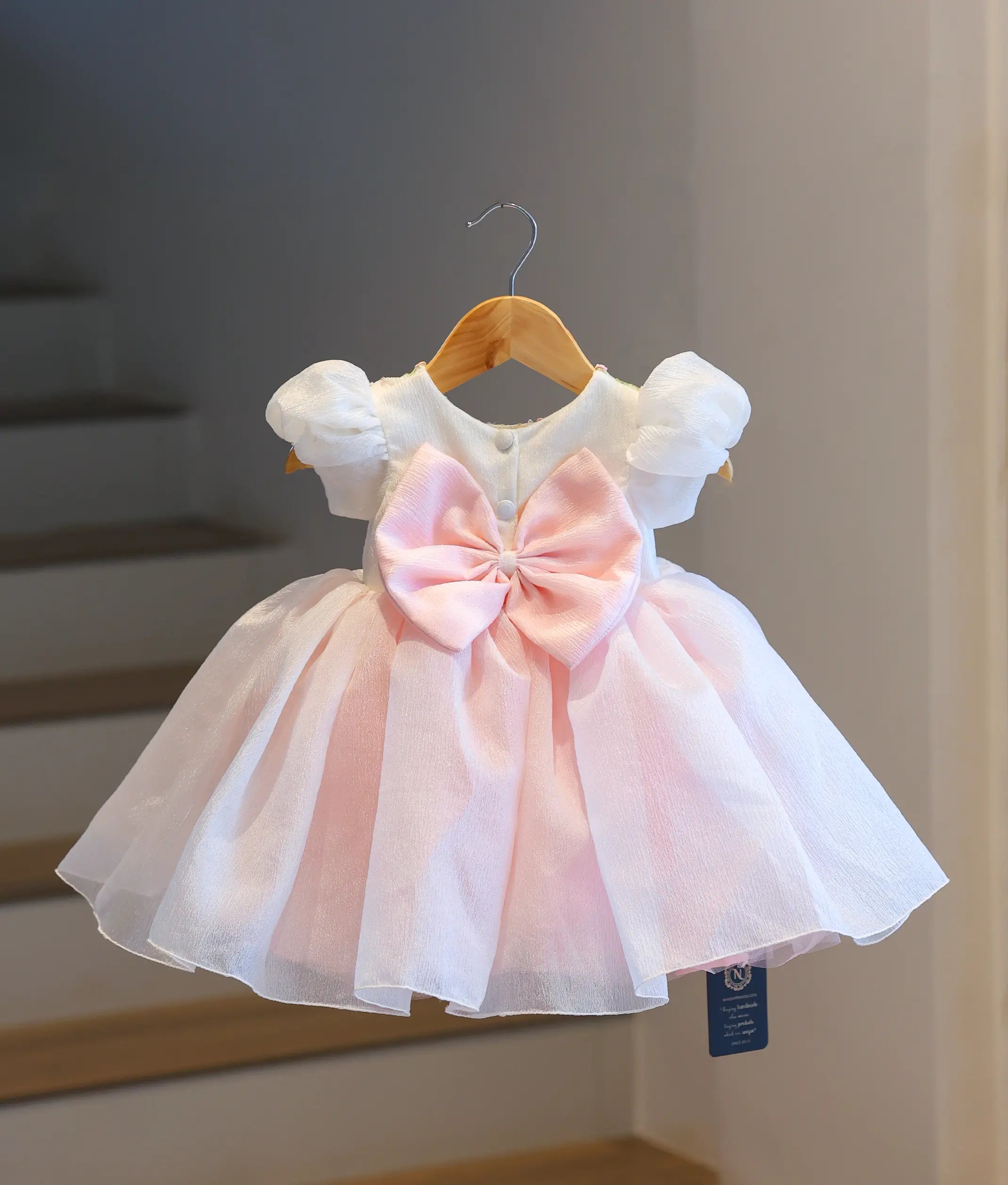 Back view of the off-white and blush pink kids party frock on a hanger, showing the round back neckline, neat button closures, and a large blush pink tie-up waist bow.