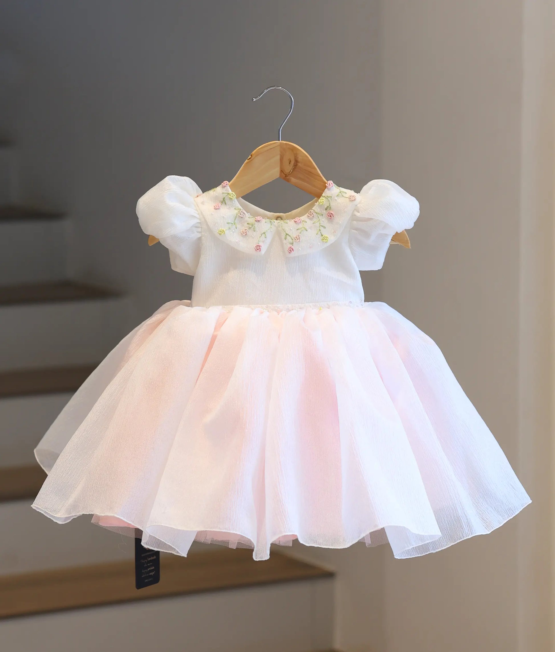 Full-length front view of the off-white and blush pink crush organza baby frock on a hanger, showcasing the floral embroidered top and the flared blush pink bottom.