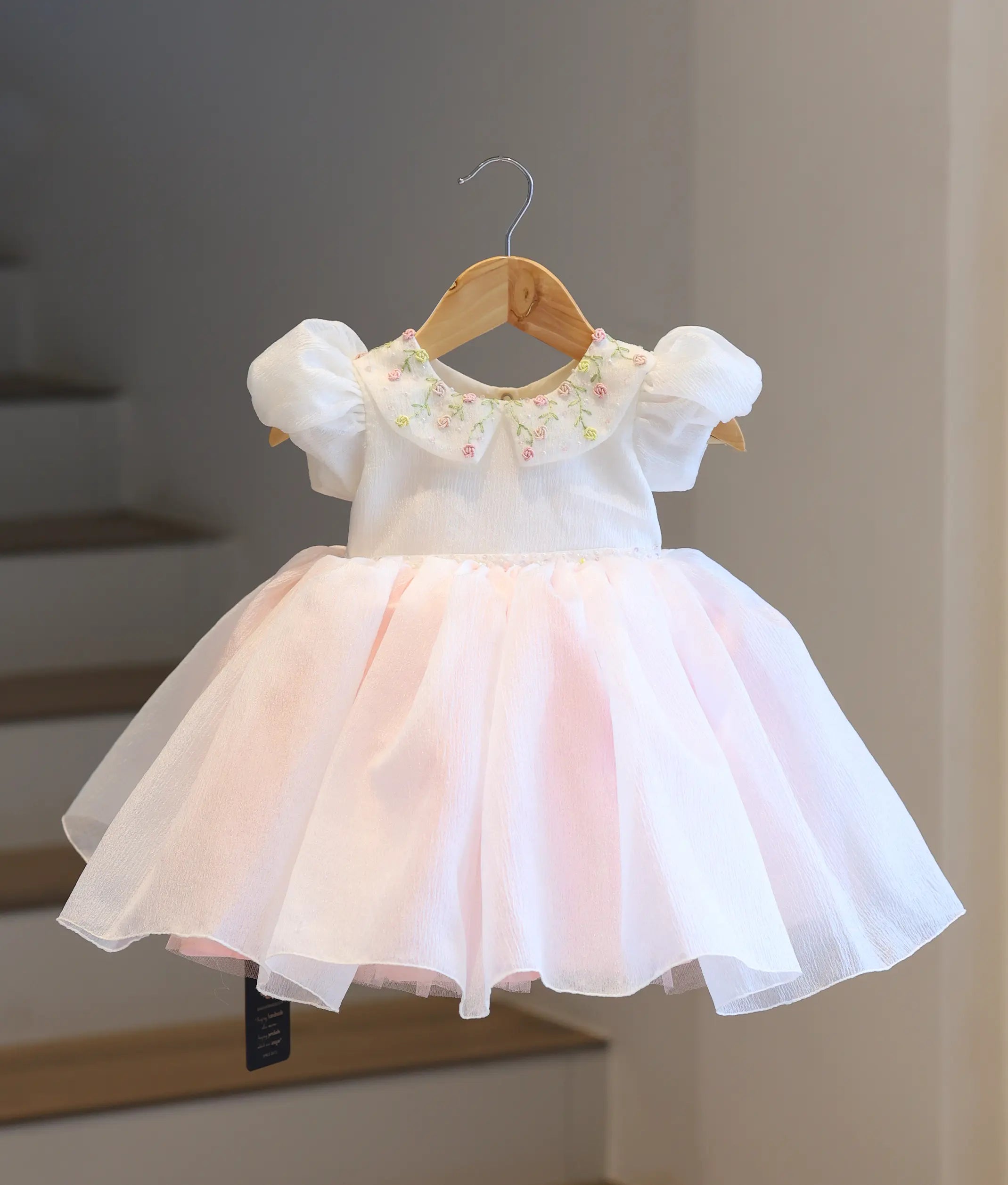 Full-length front view of the off-white and blush pink crush organza baby frock on a hanger, showcasing the floral embroidered top and the flared blush pink bottom.