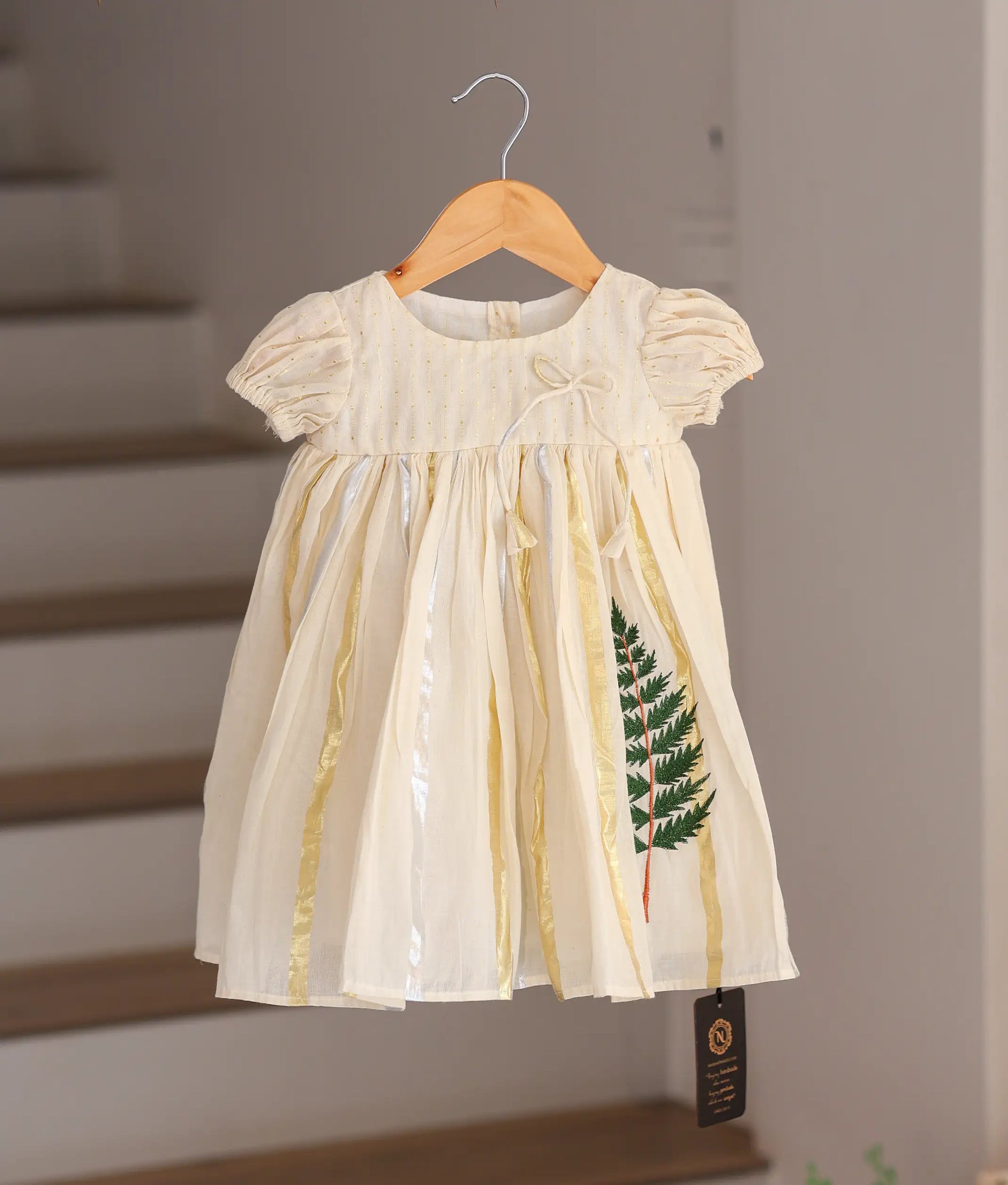Front view of a traditional off-white cotton baby frock with gold tissue stripes and green leaf embroidery