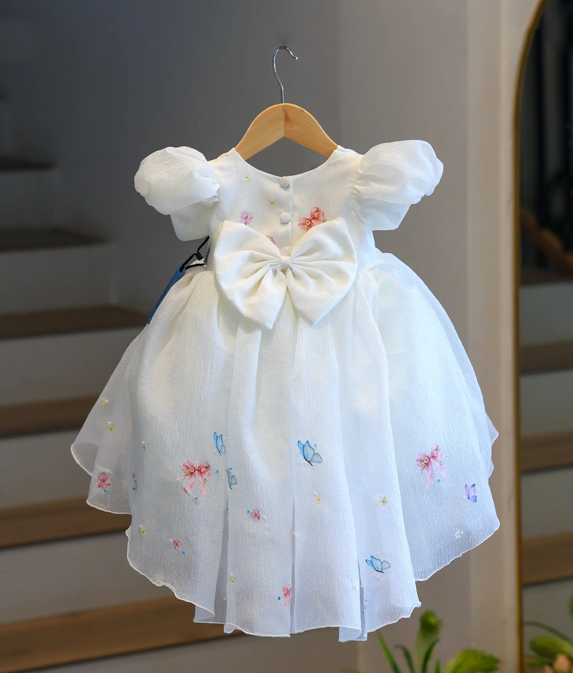 Back view of the off-white crush organza high-low frock, showcasing the round back neck with button closures, embroidered butterflies, and a large, puffy matching off-white bow at the waist.