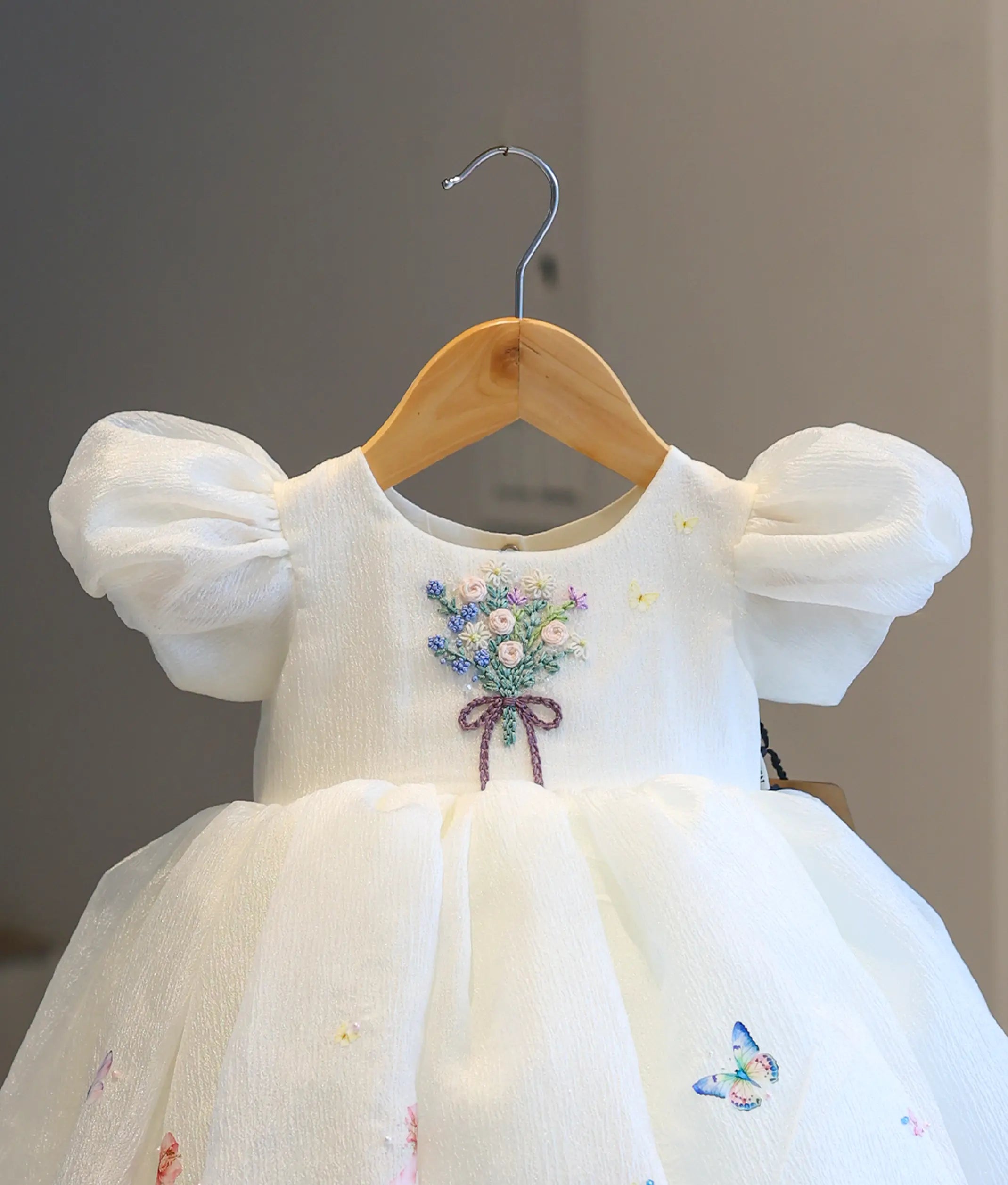 Close-up detail of the off-white dress bodice, showcasing the hand-embroidered floral bouquet with a purple bow, delicate butterfly details, and sheer puffed sleeves.