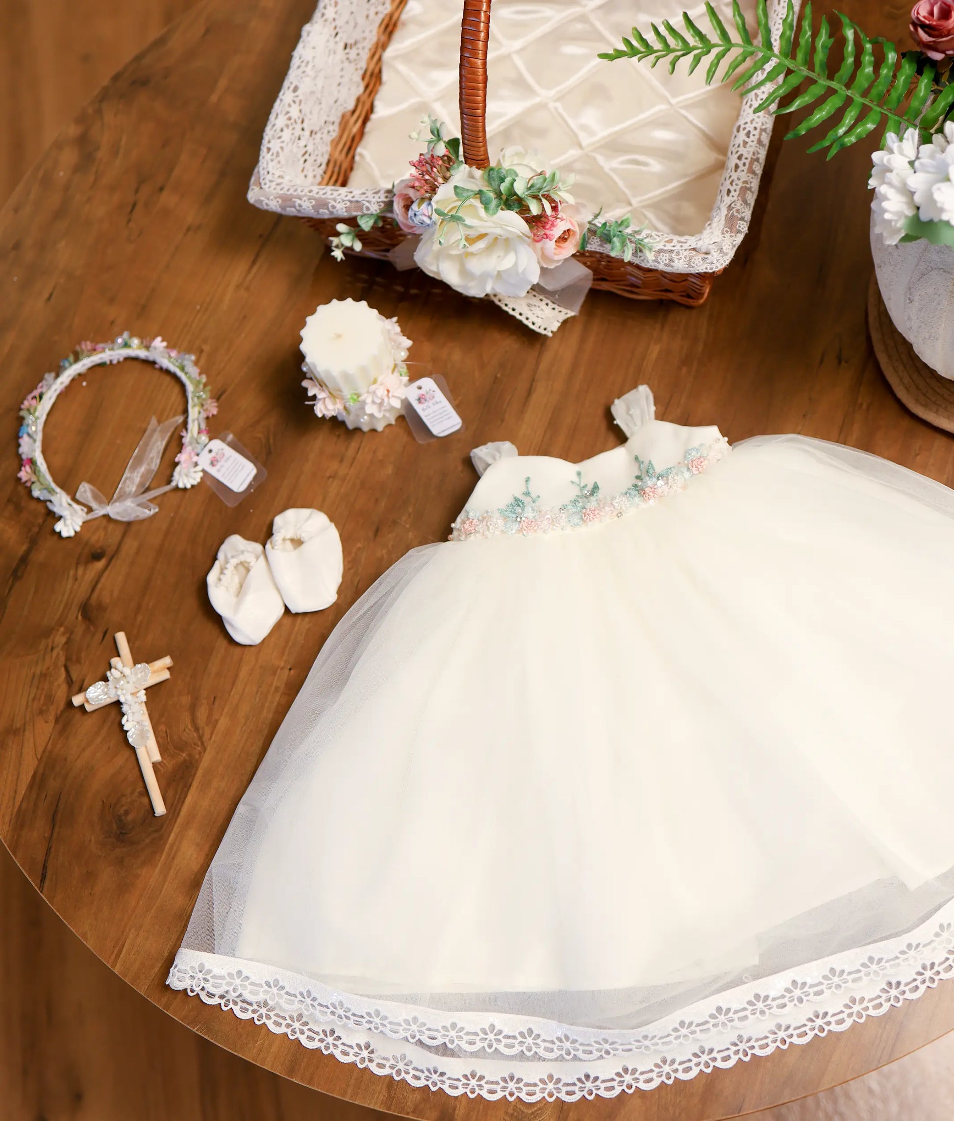 Off-white baby girl baptism set featuring tulle dress with floral waistline embroidery, booties, floral crown headband, wooden cross, decorated candle, and lace-lined basket.