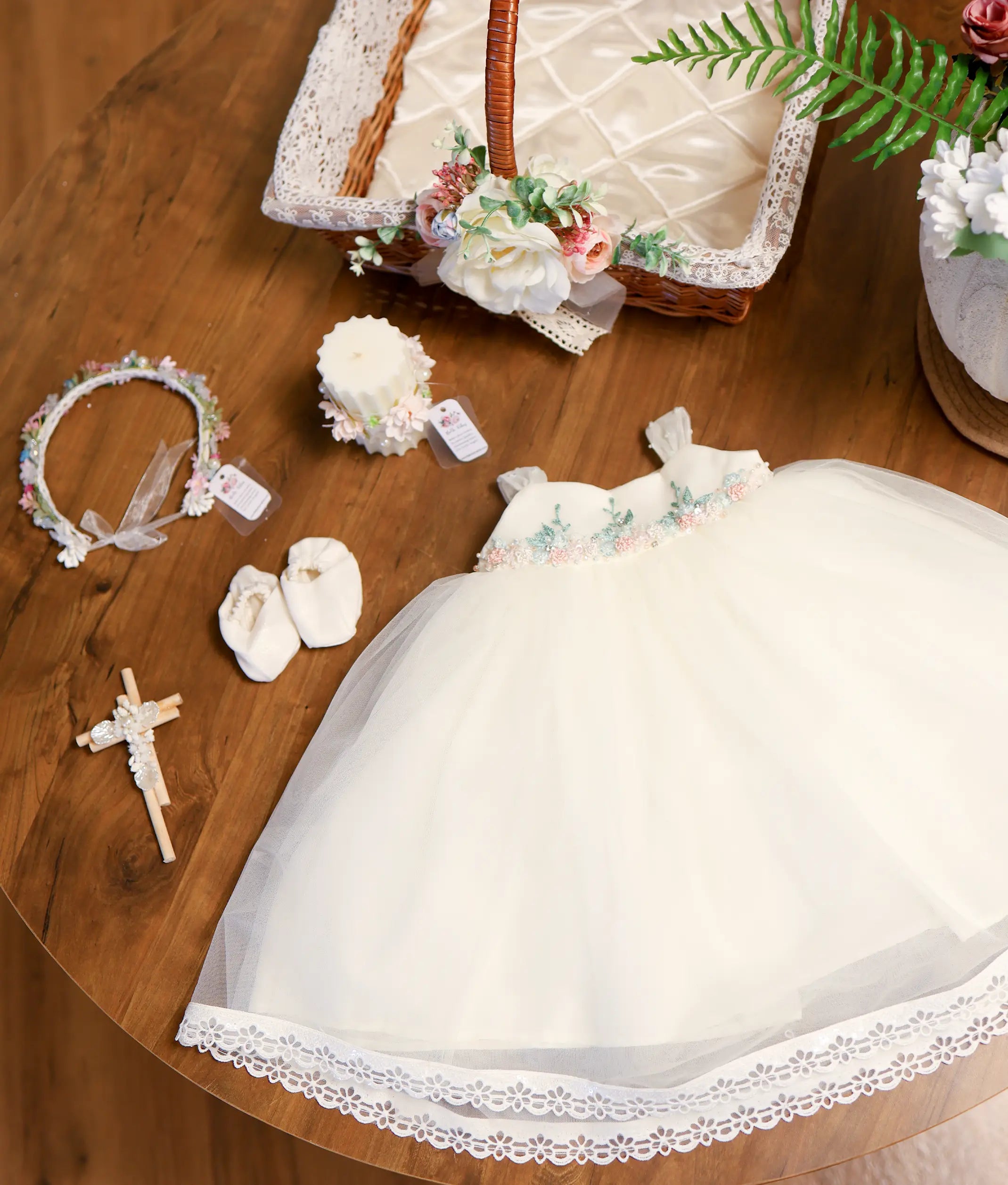 Off-white baby girl baptism set featuring tulle dress with floral waistline embroidery, booties, floral crown headband, wooden cross, decorated candle, and lace-lined basket.