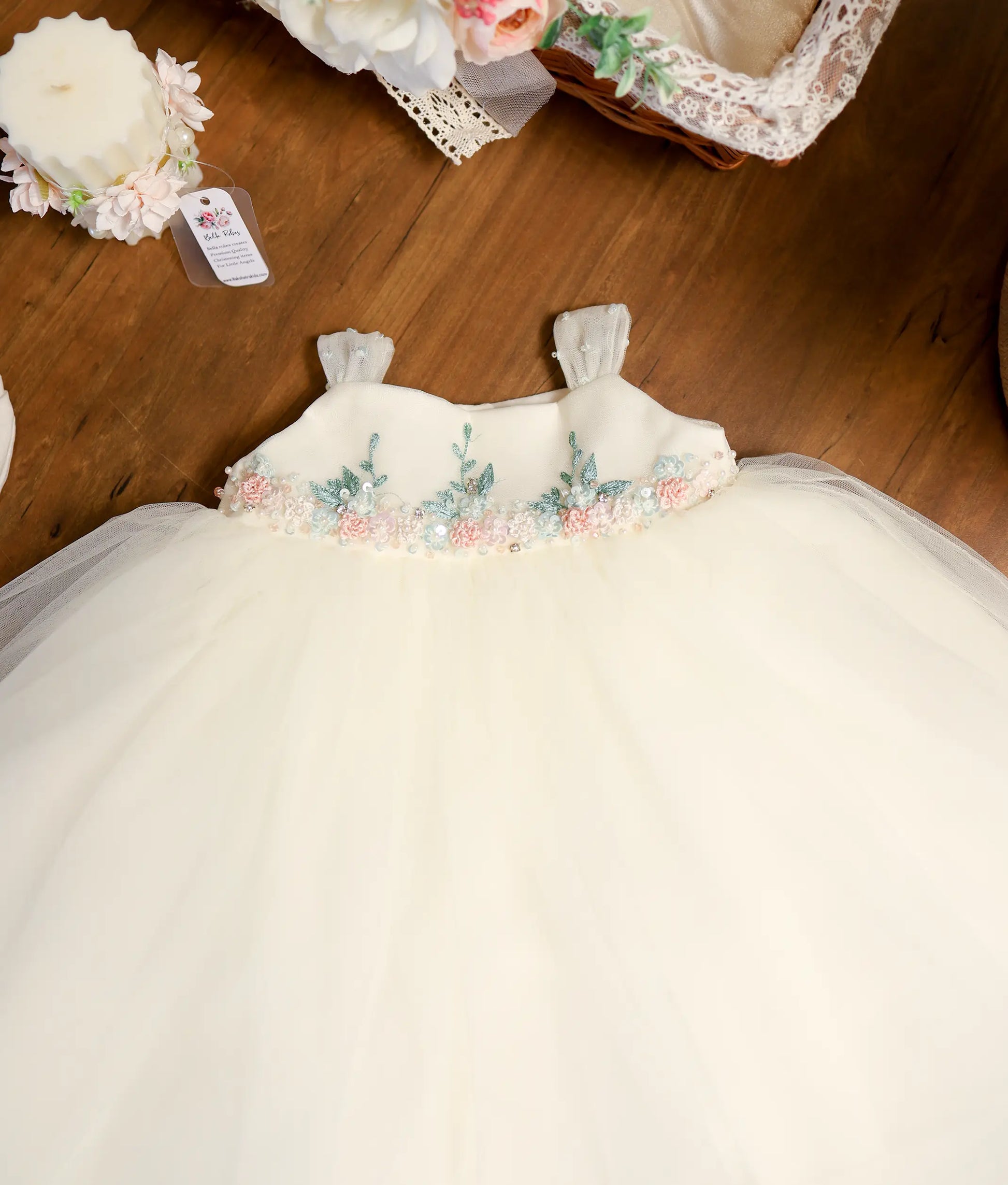 Close-up of off-white baptism dress showing pastel floral handwork embroidery with pink and mint green accents on the waistline.