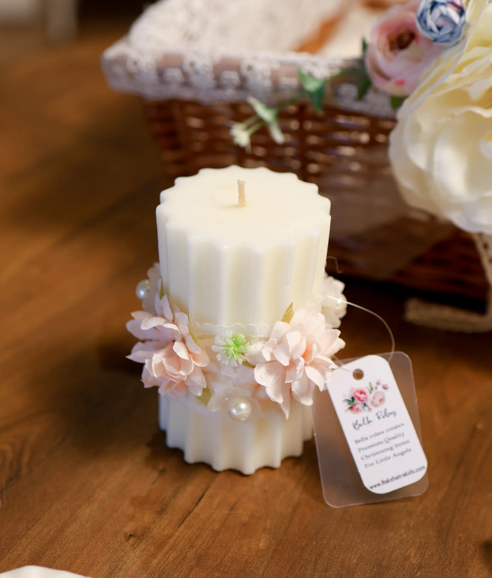 Textured off-white pillar baptism candle decorated with pink fabric flowers, green accents, lace wrap, and pearl embellishments.