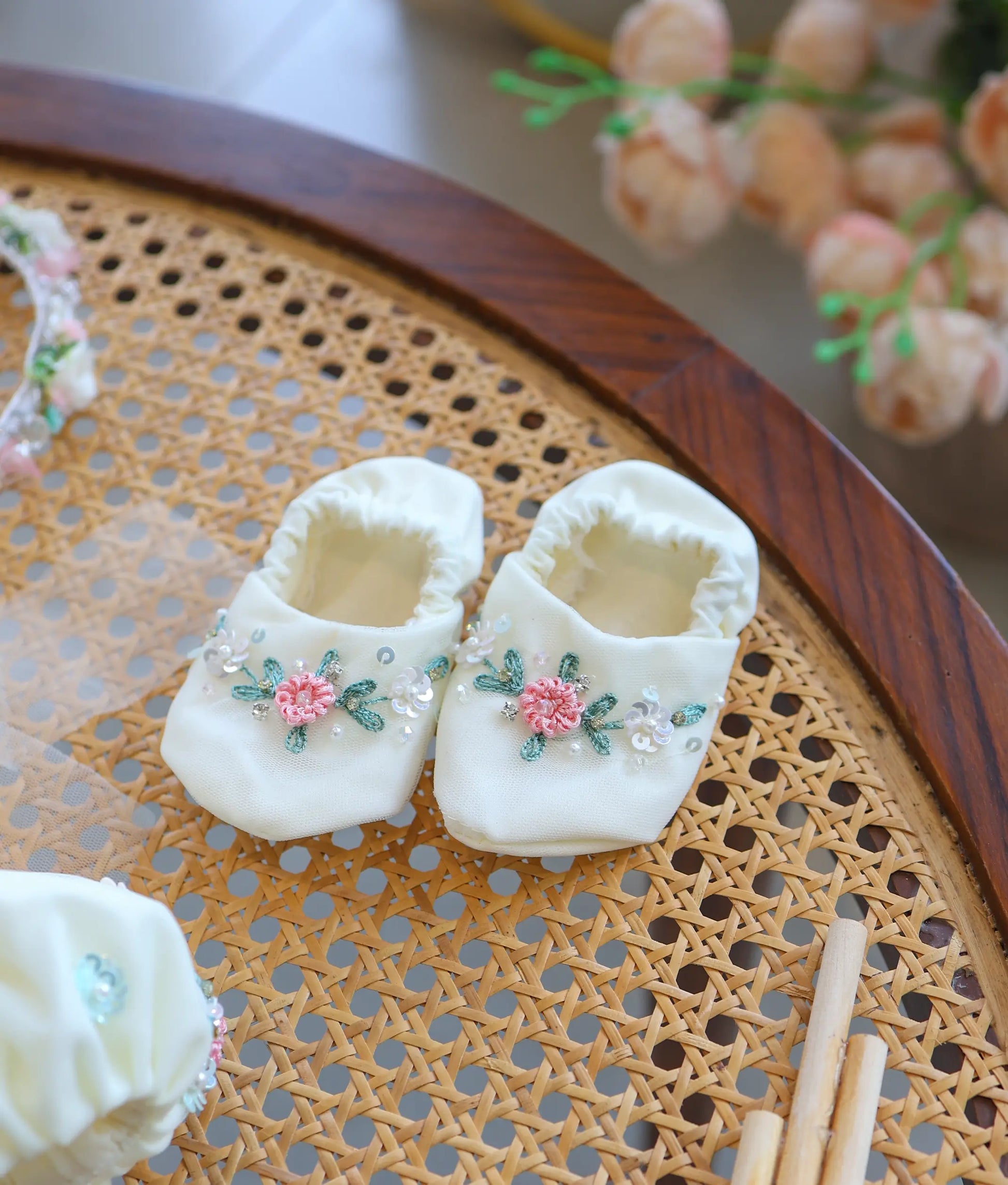 Pair of off-white soft fabric baby booties featuring pink floral handwork embroidery and sequin details.