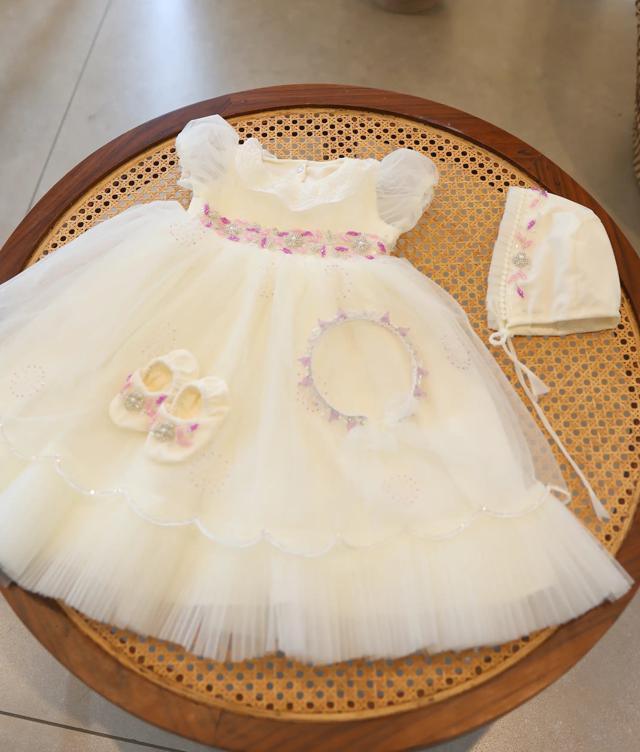 Full off-white baby girl baptism set with pink and silver waist embroidery, matching bonnet cap, booties, and headband on a cane table.