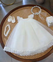Complete off-white baby girl baptism set showing tulle frock with lace hem, matching booties, floral crown, elastic headband, and customized candle on a cane table.