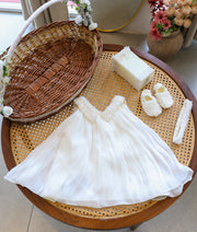 Complete off-white baby girl baptism set featuring pleated organza frock with pearl collar, pearl booties, embellished candle, and elastic headband on a cane table.
