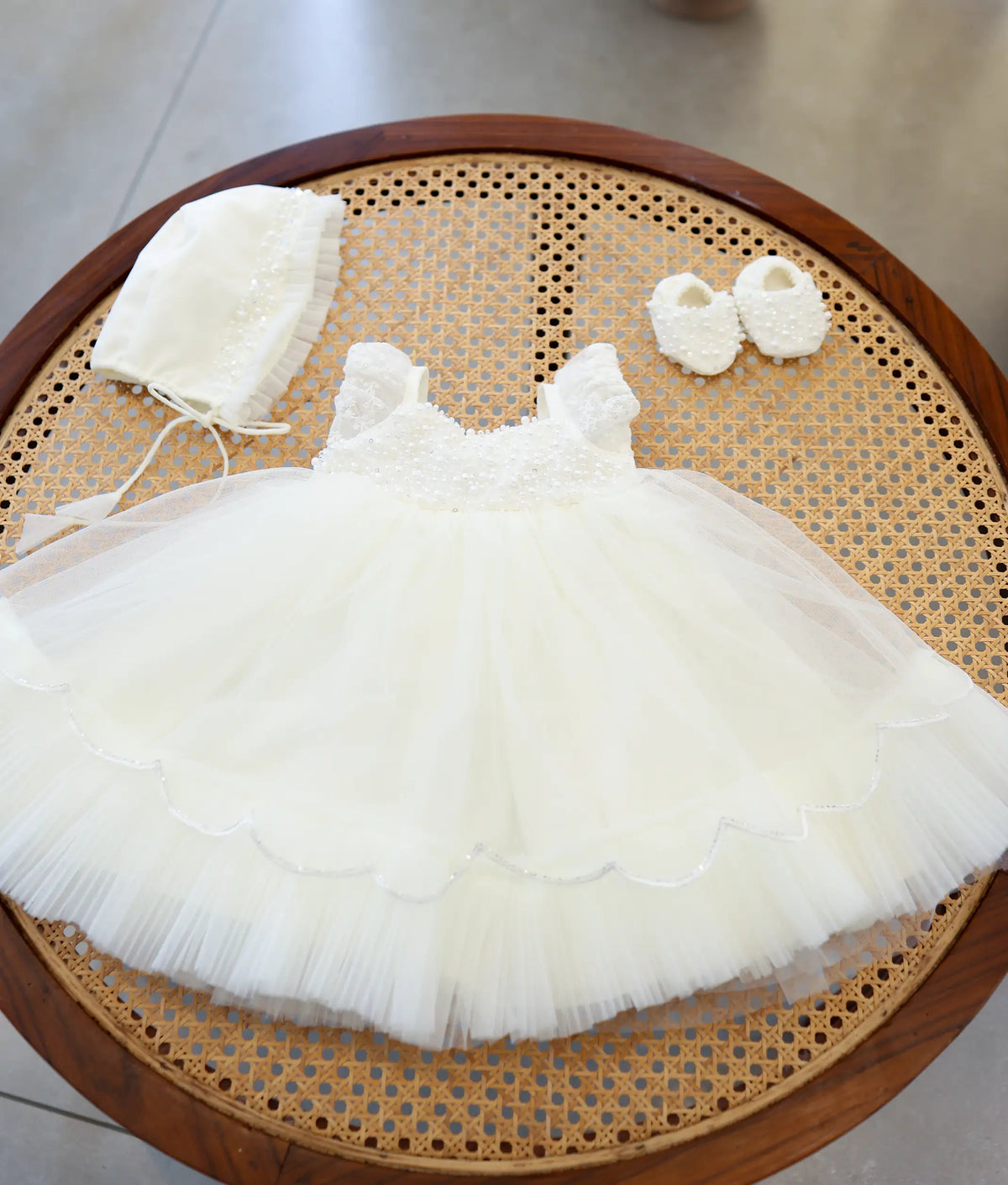 Complete off-white handwork baptism set for baby girl featuring pearl-embellished net frock, matching bonnet cap, and handworked booties on a woven cane table