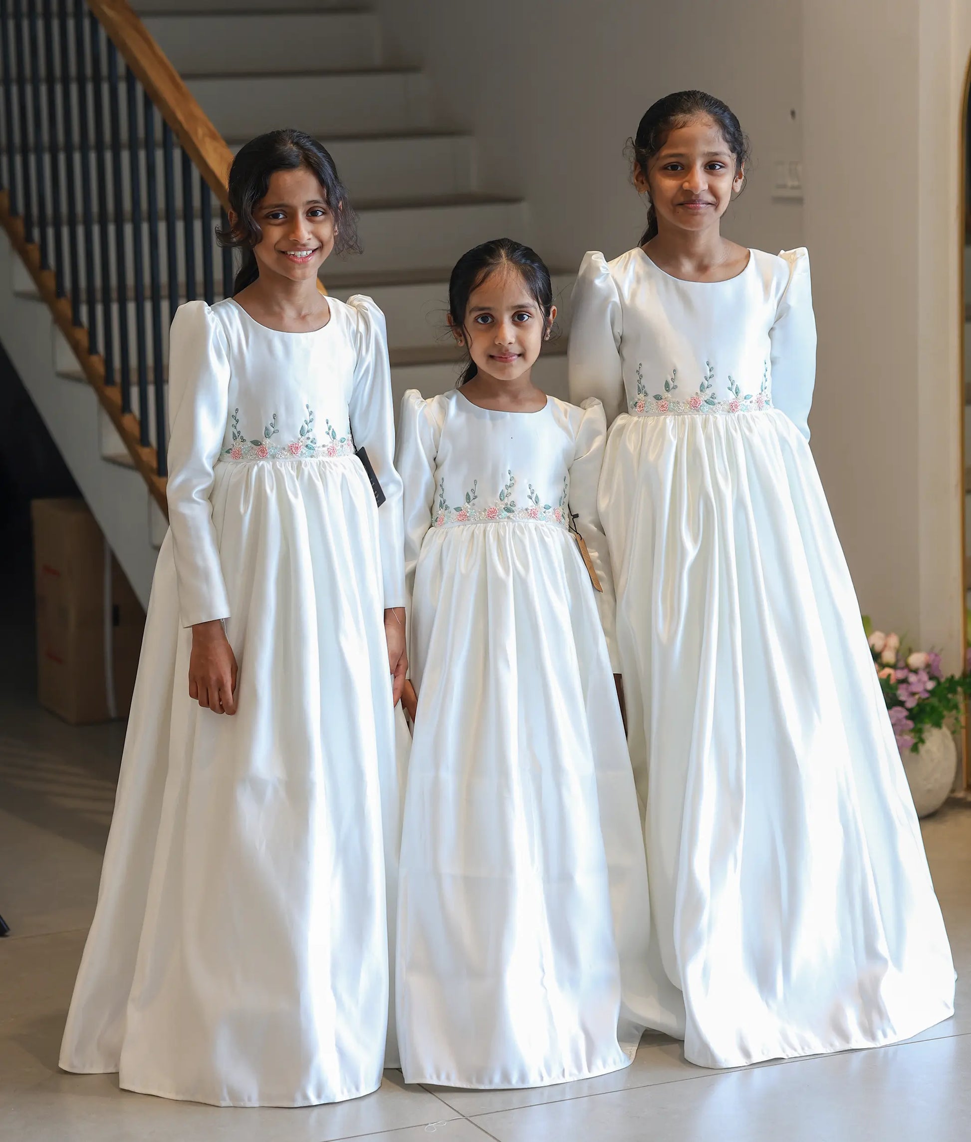 Three young girls smiling while wearing matching long-sleeve off-white satin Holy Communion gowns with pastel floral waist embroidery.