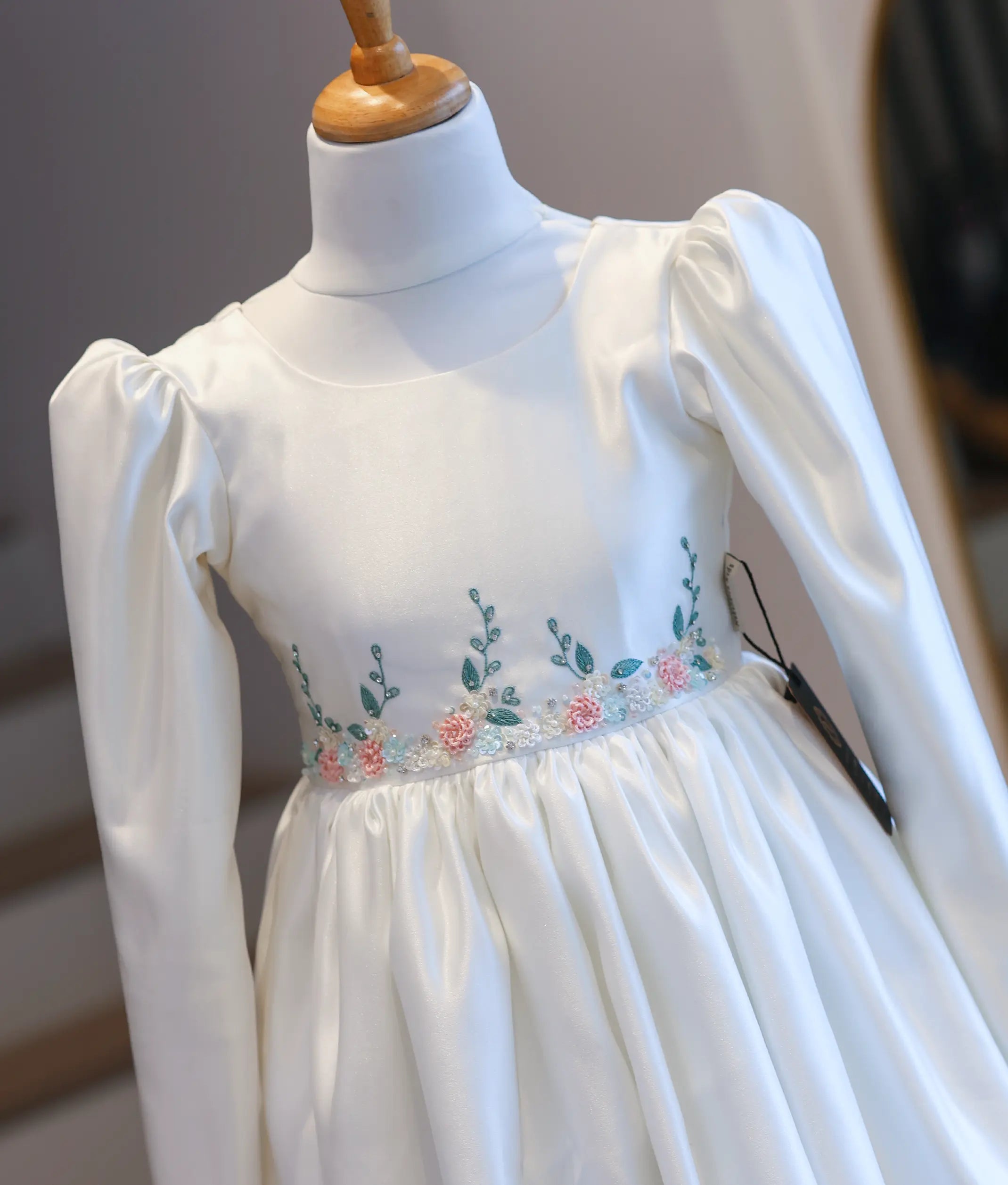 Close-up display of the communion dress bodice on a mannequin, showcasing the puff sleeves and the detailed pastel pink, blue, and green floral embroidery at the waist.