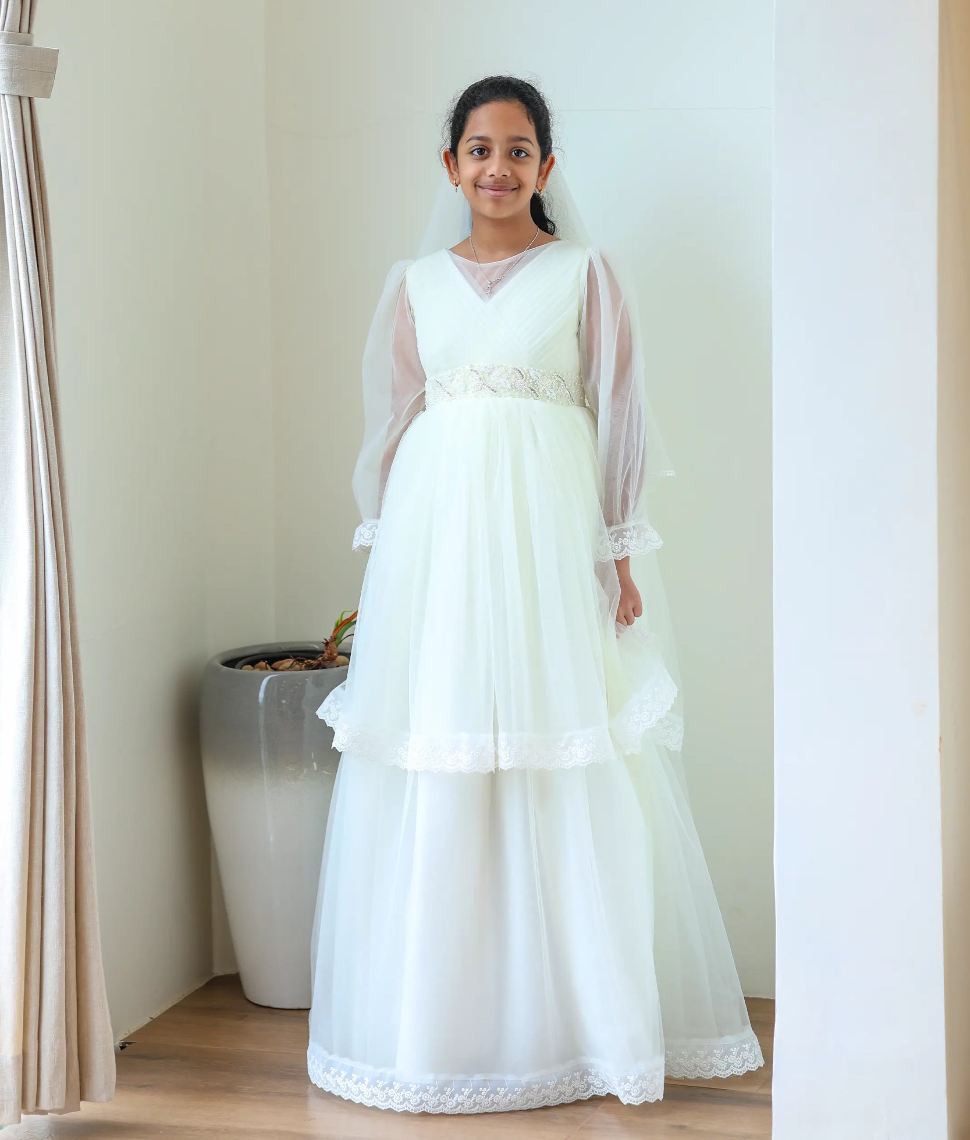 A girl smiling while wearing the Nakshatra Kids elegant off-white Holy Communion gown, complete with a matching sheer white net head veil.
