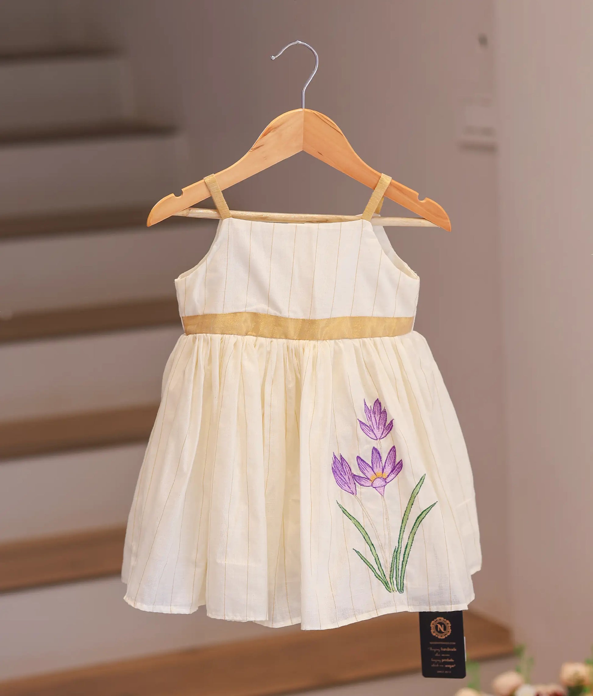 Front view of an off-white kasavu frock for girls featuring gold straps and a pleated bottom.