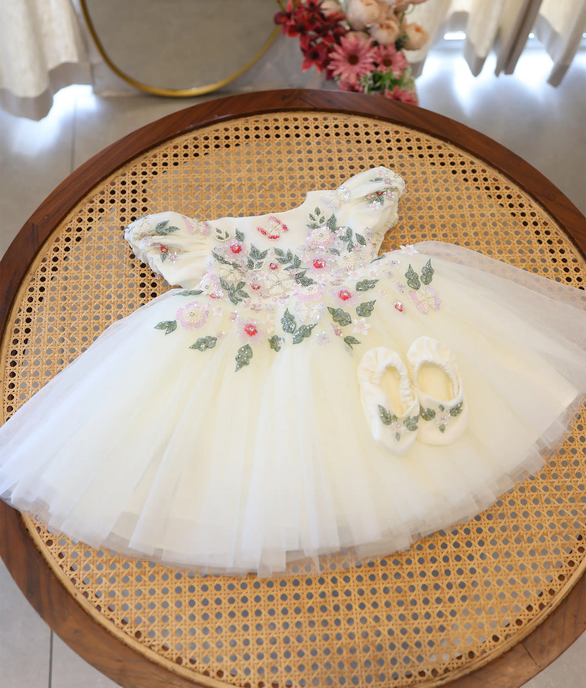 Full view of off-white tulle net christening gown with handworked floral yoke and matching booties set on rattan table.