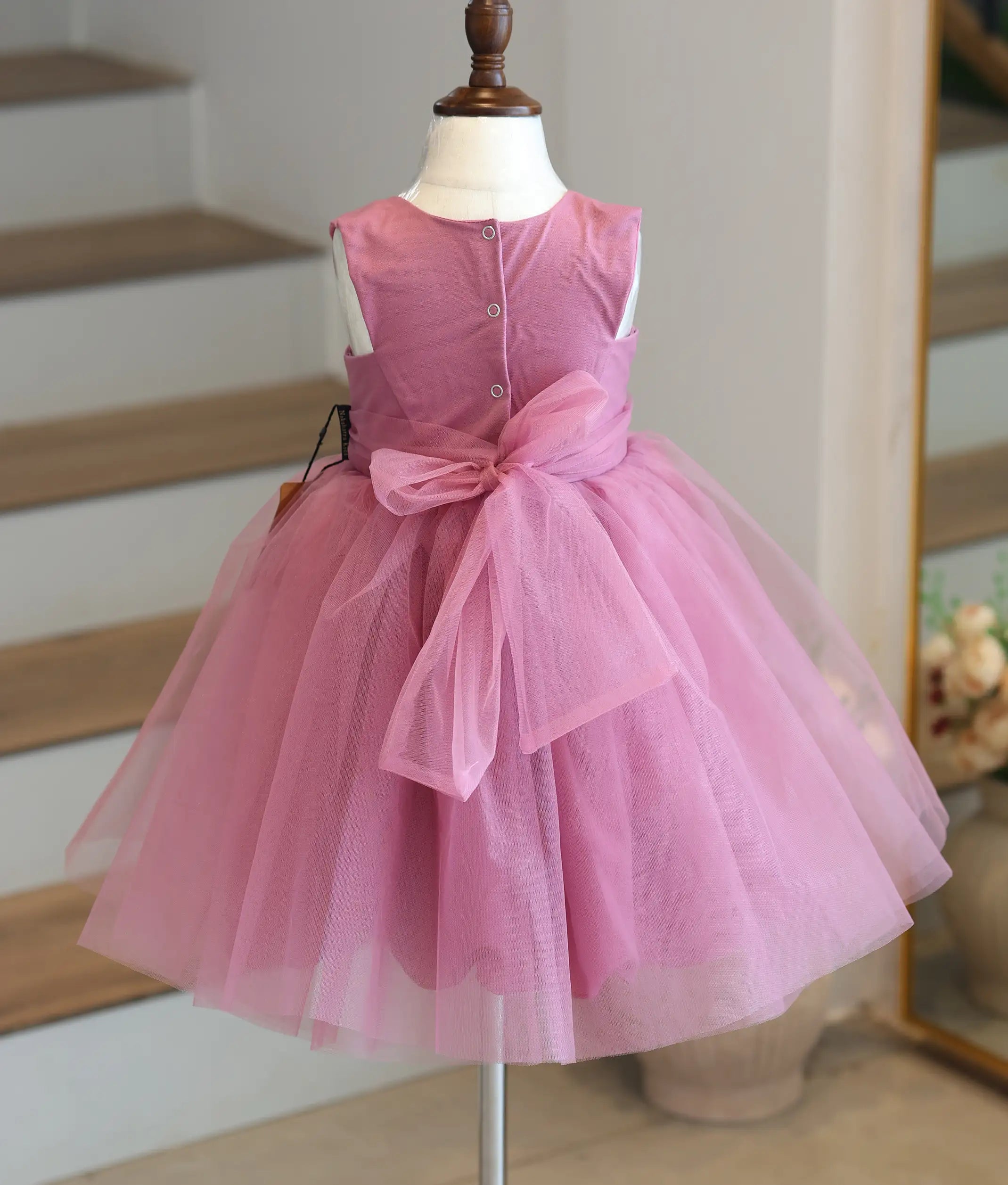 Back view of an onion-pink below-knee-length girls' frock with a large net bow and button closures.