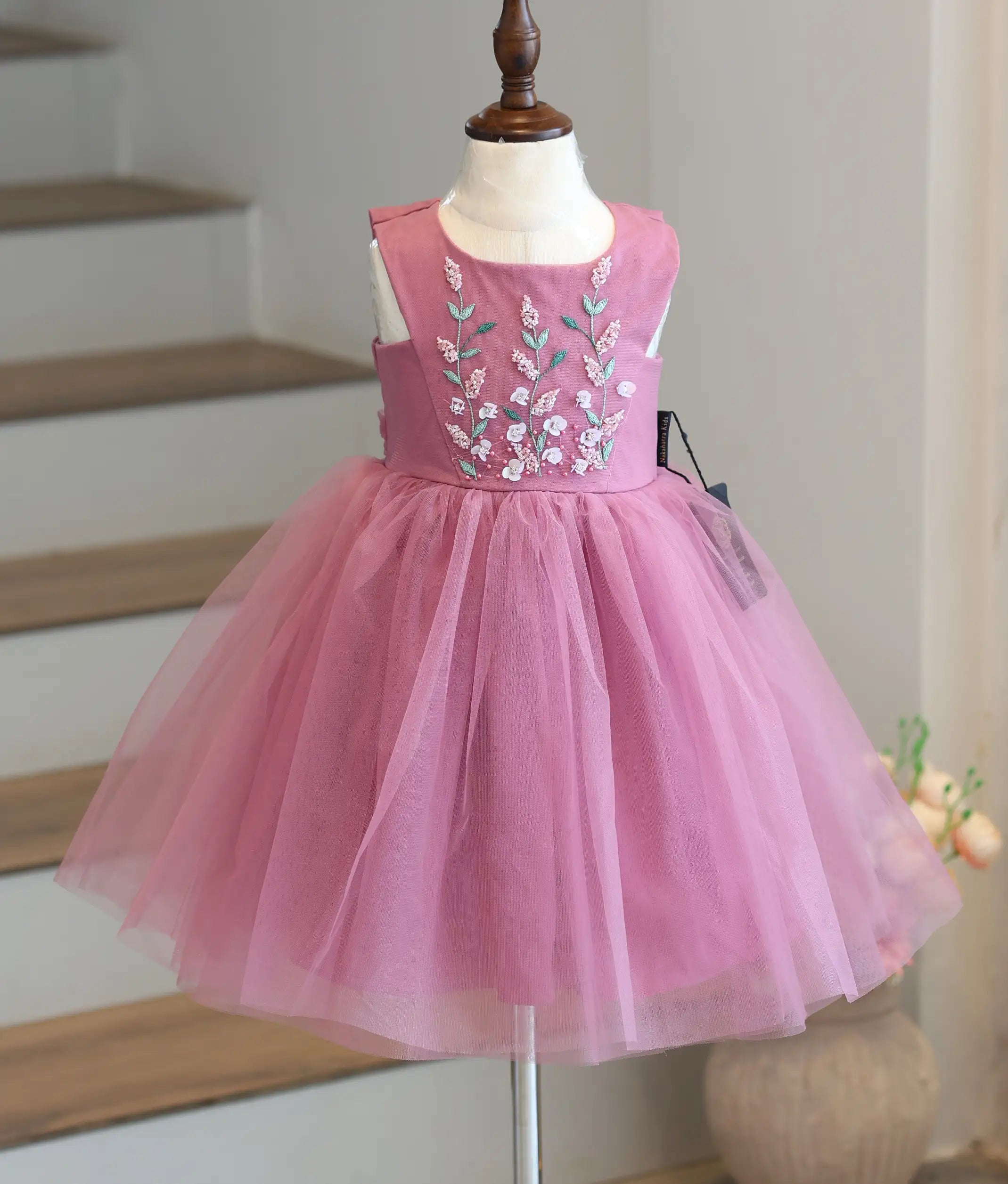 Front view of a sleeveless onion-pink girls' frock with floral handwork on the yoke and a voluminous net skirt falling below the knee.