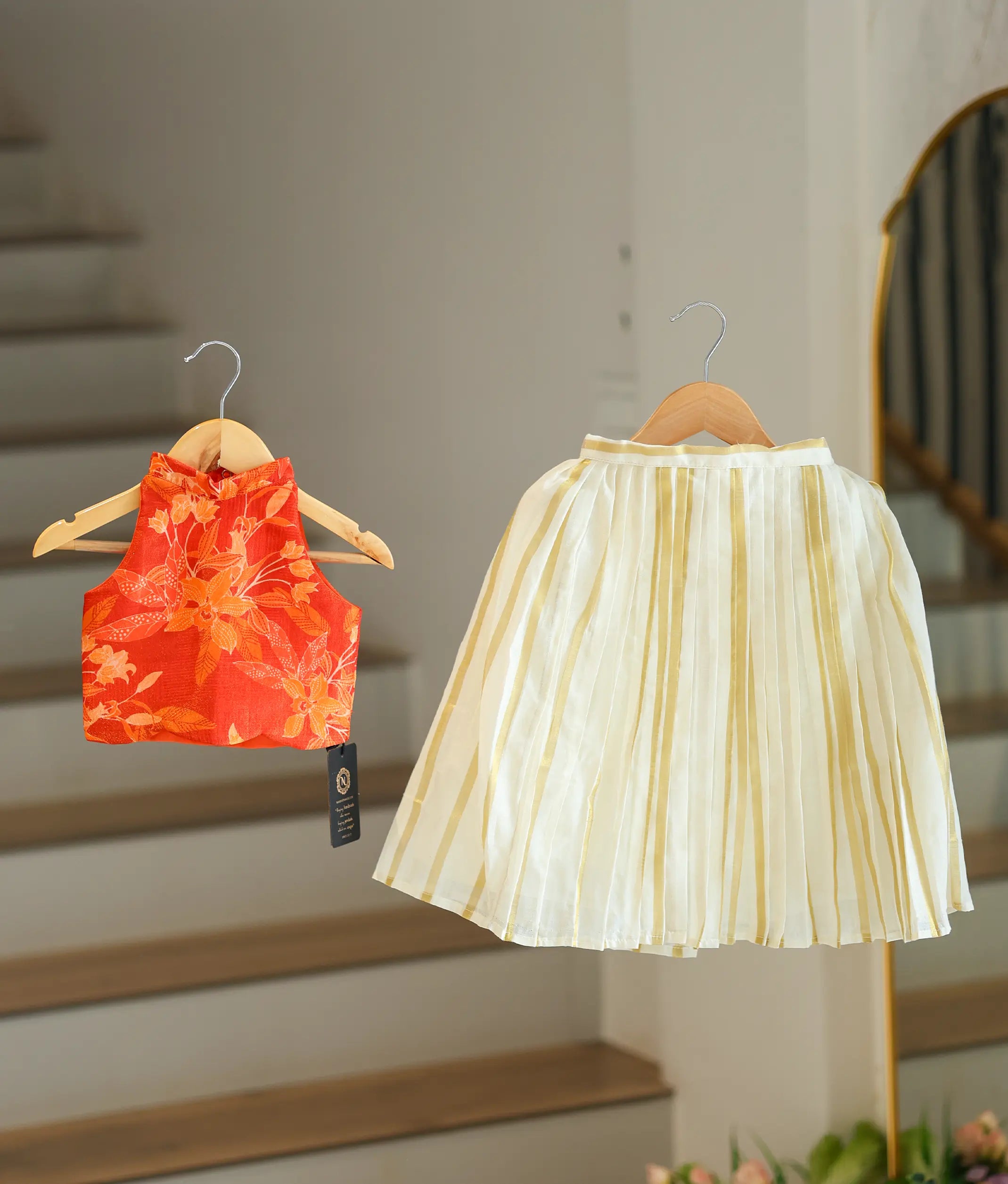 Front view of a girls' ethnic set featuring an orange floral sleeveless top and a pleated with vertical kasavu stripes.
