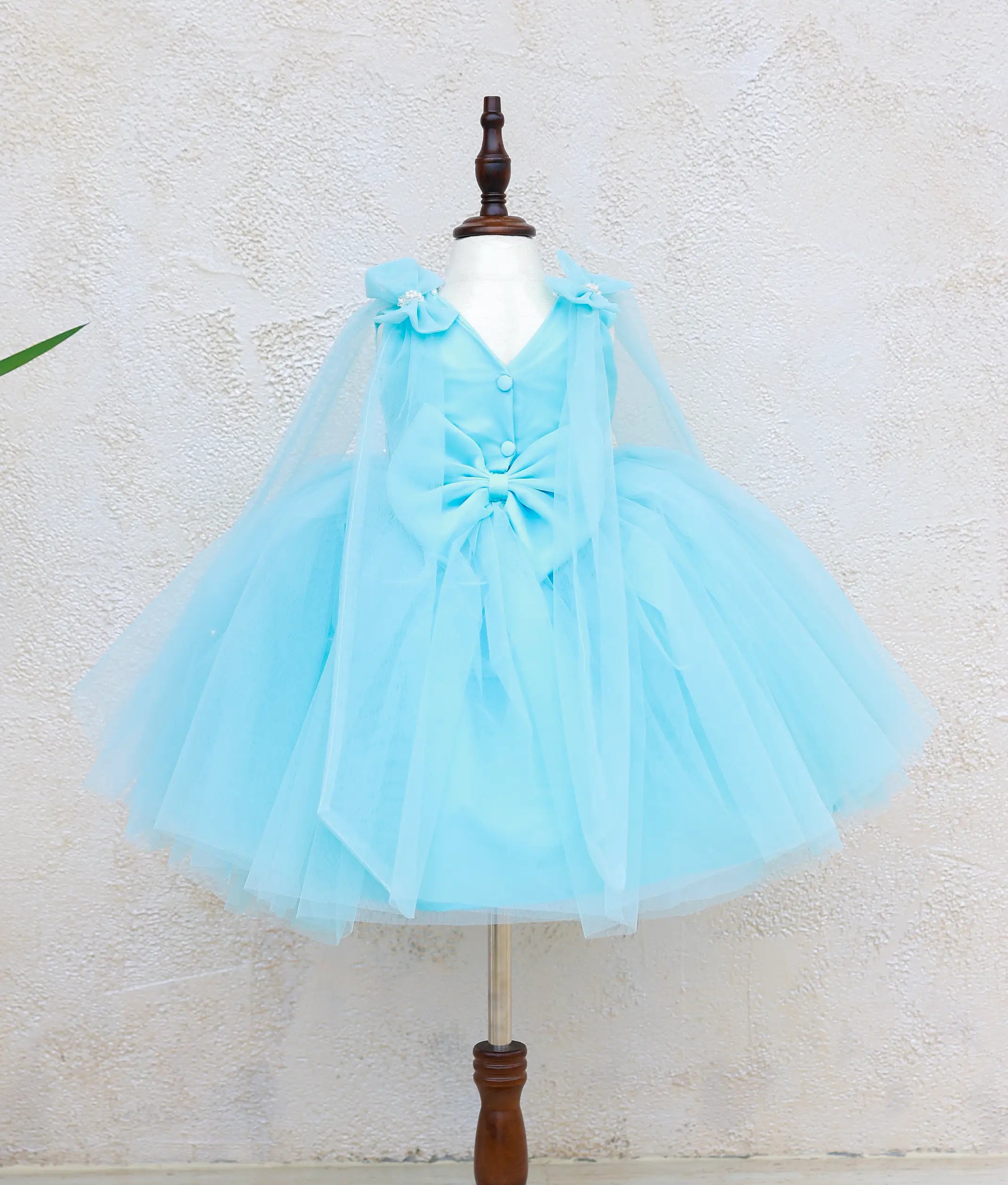 Back view of sky blue Frozen Elsa theme dress for baby girl with V-neck, decorative buttons, oversized bow, detachable tulle cape, and layered tulle skirt.