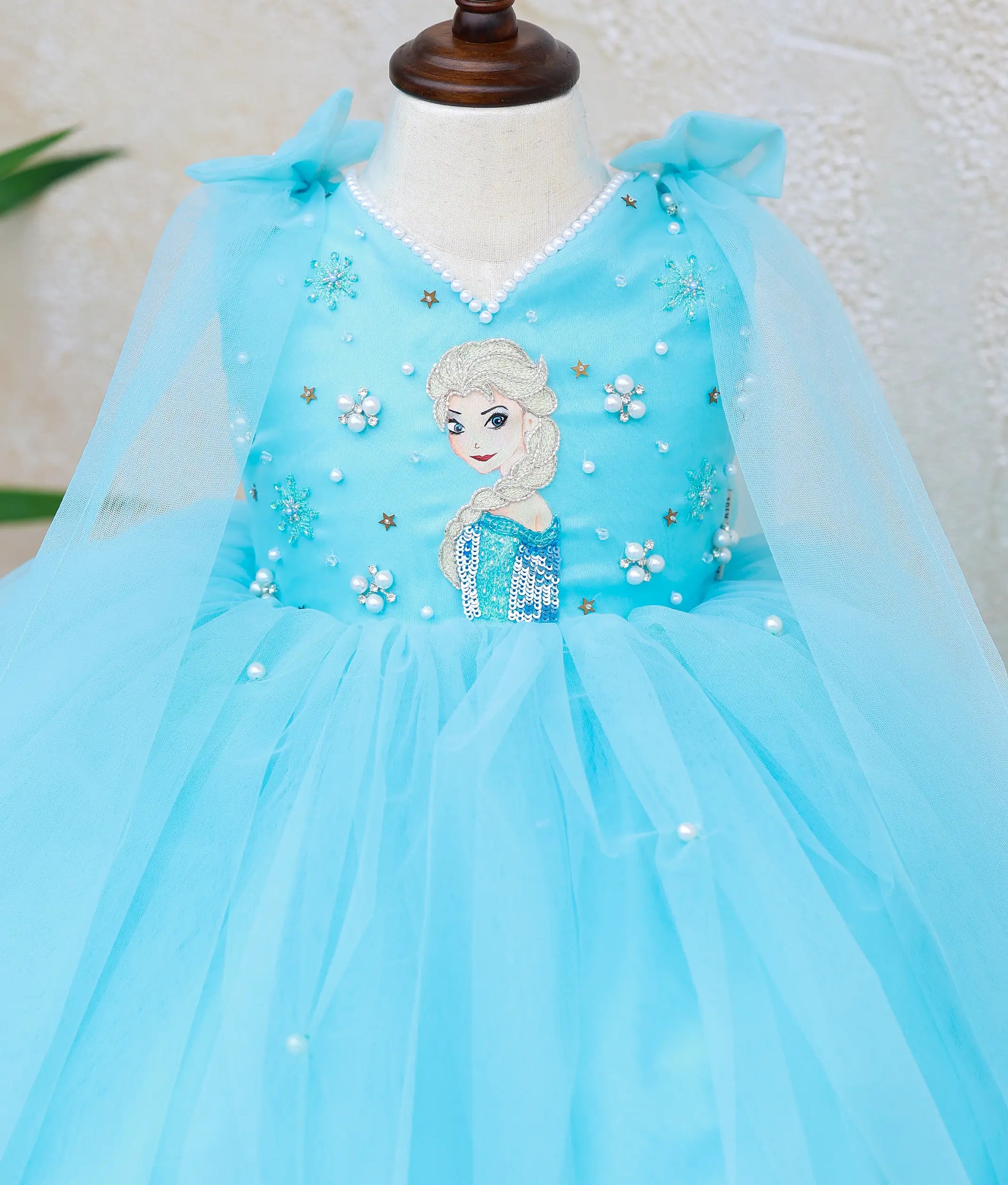 Close-up of sky blue Frozen Elsa theme baby girl dress featuring handworked Elsa embroidery, pearl bead detailing, V-neck, and sheer tulle cape.
