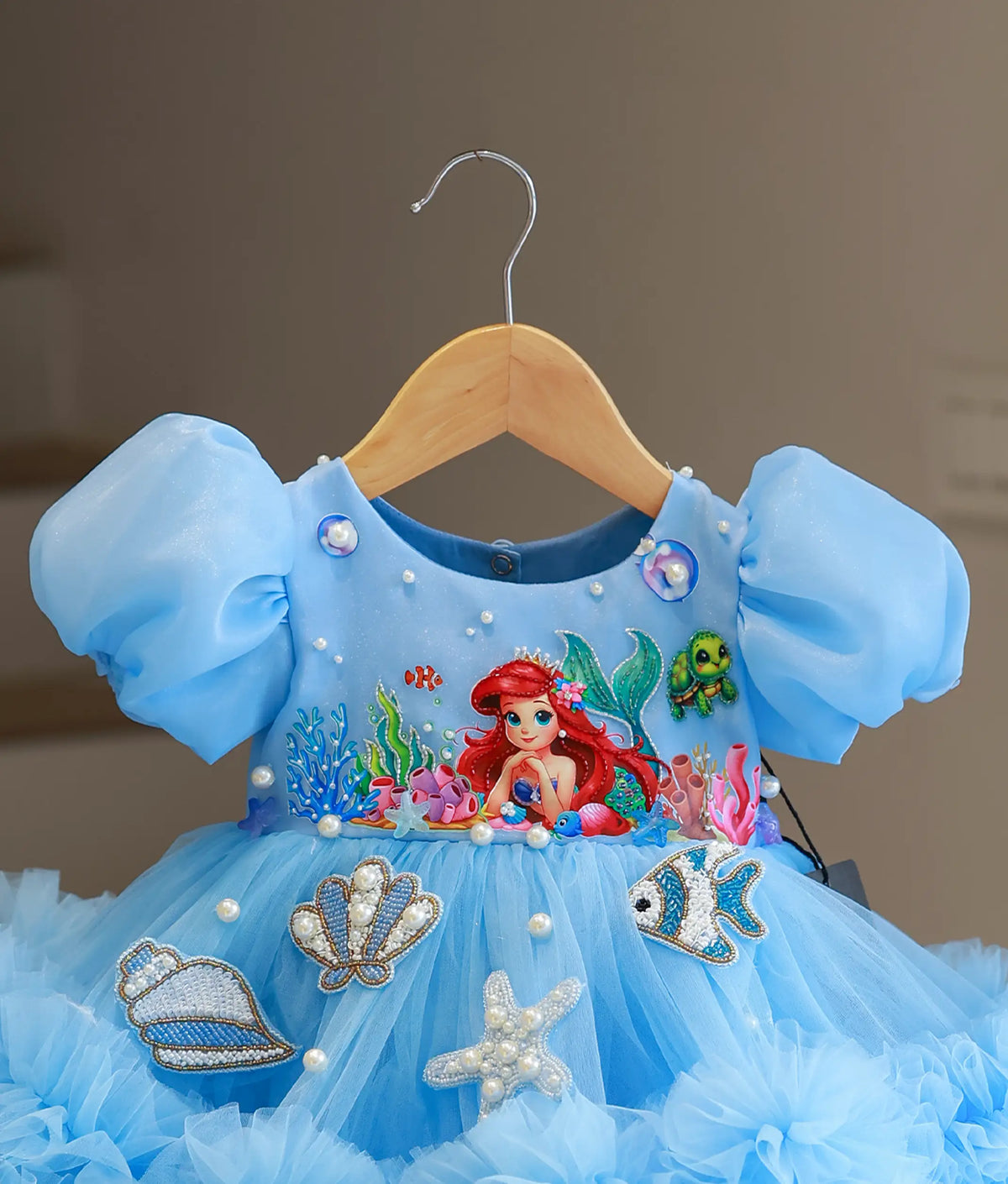 A light blue toddler girl's party dress featuring Ariel from The Little Mermaid, hanging on a wooden hanger against a neutral indoor background. The dress has puffy sleeves, a bodice with embroidered sea creatures, and a layered tulle skirt decorated with sequined shells, a starfish, and faux pearls.
