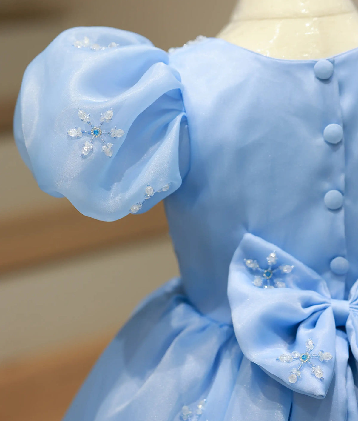 Close-up back view of sky blue gown showing button closure, puffed sleeve with snowflake handwork, and waist bow.