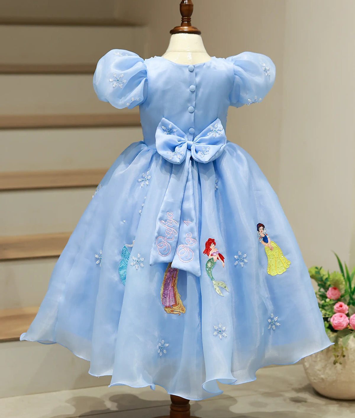 Back view of sky blue organza gown showing waist bow with personalized name embroidery and princess skirt border.