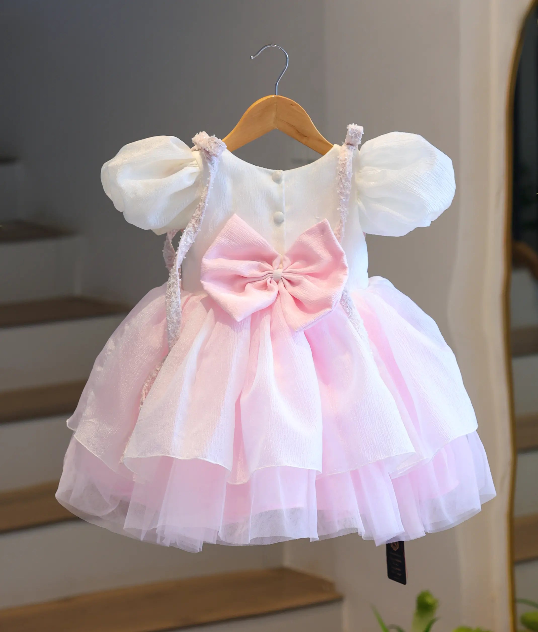 Back view of the white and baby pink strawberry theme crush organza party frock on a hanger, featuring the button closures and a large pink waist bow.