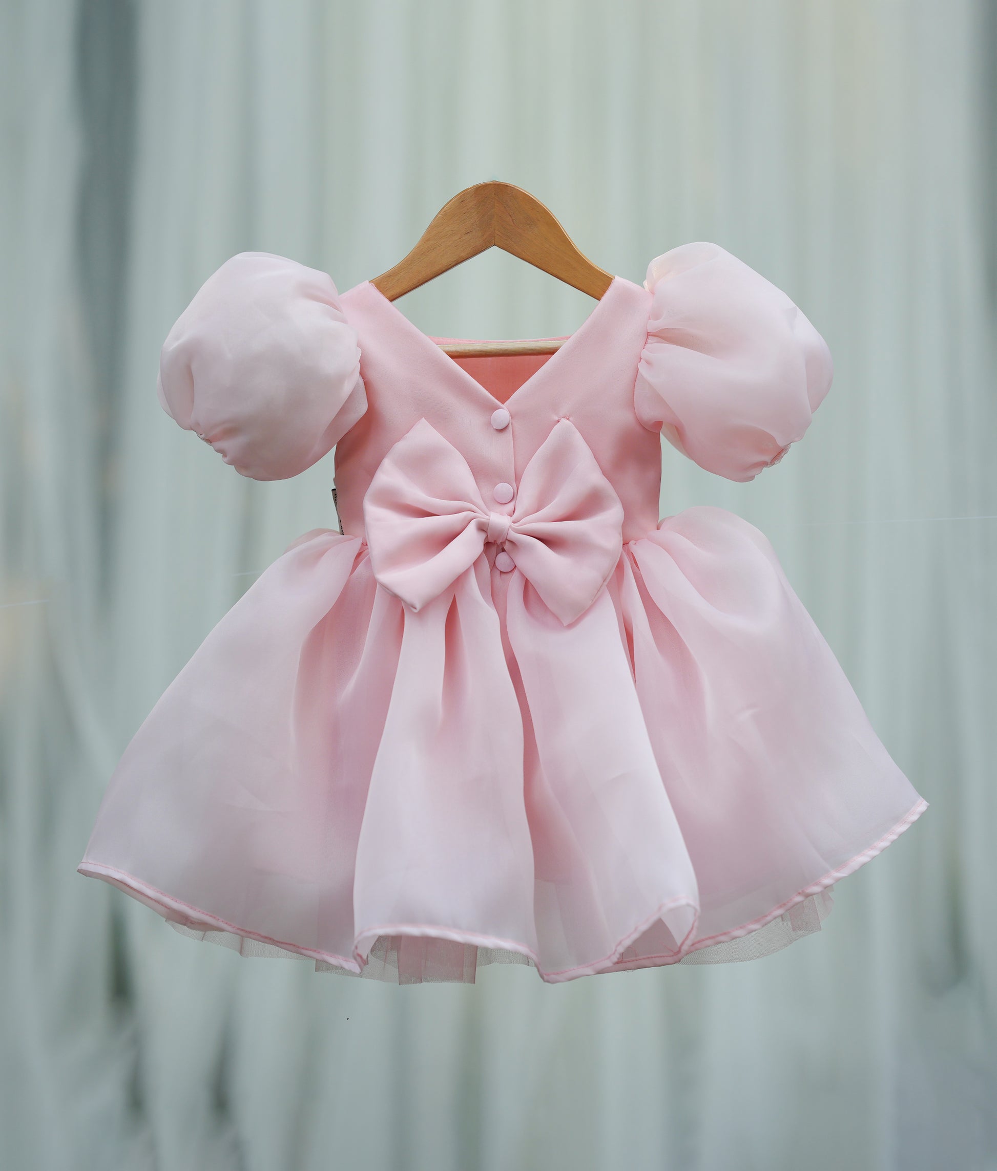 Back view of the powder peach organza frock featuring a lace-detailed V-neck, press button, and decorative bow