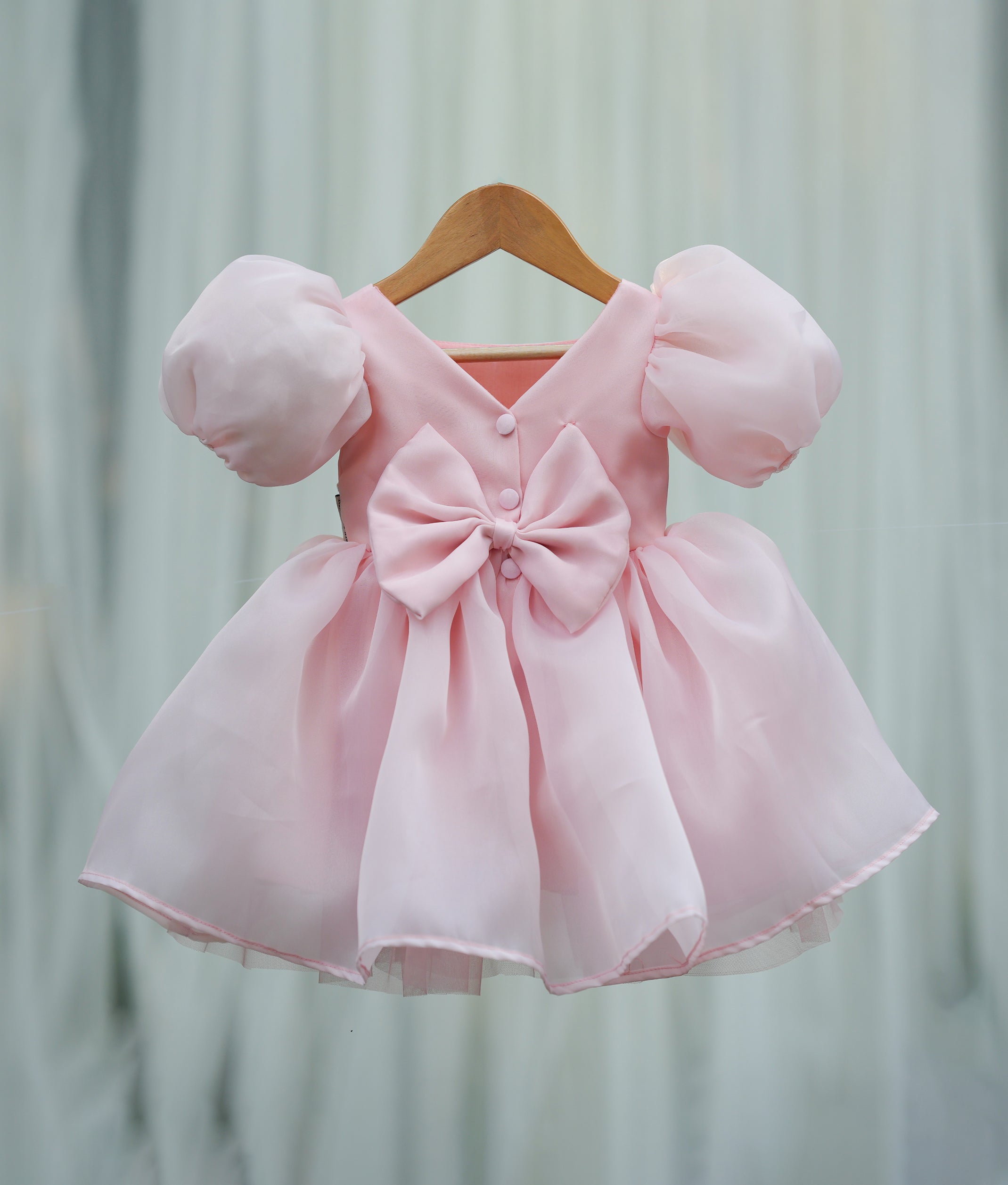 Back view of the powder peach organza frock featuring a lace-detailed V-neck, press button, and decorative bow