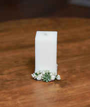 Front view of a white baptism candle decorated with white flowers, green leaves, and a white satin ribbon