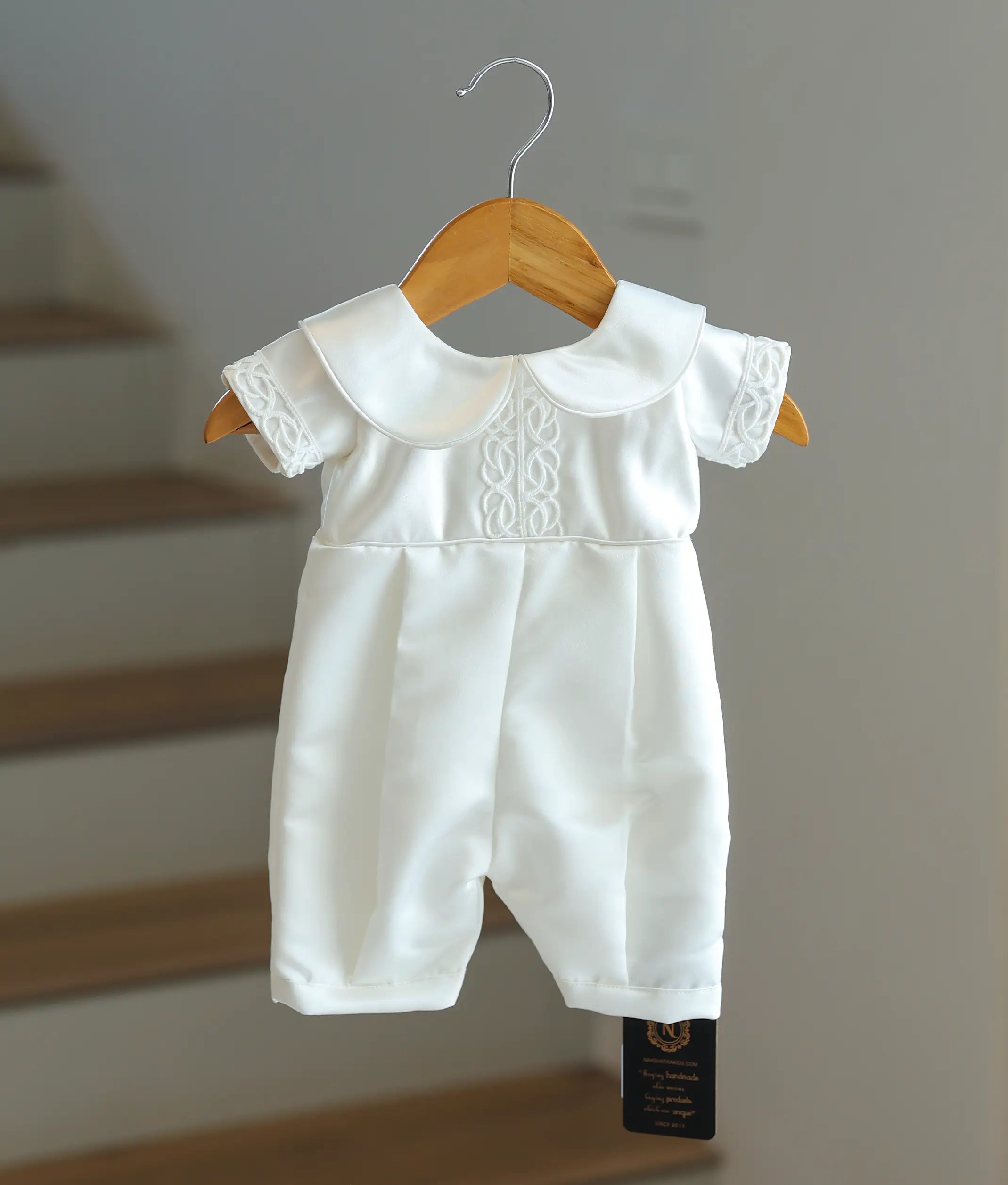 Front view of an elegant all-white satin baptism romper with pleats and a round collar.