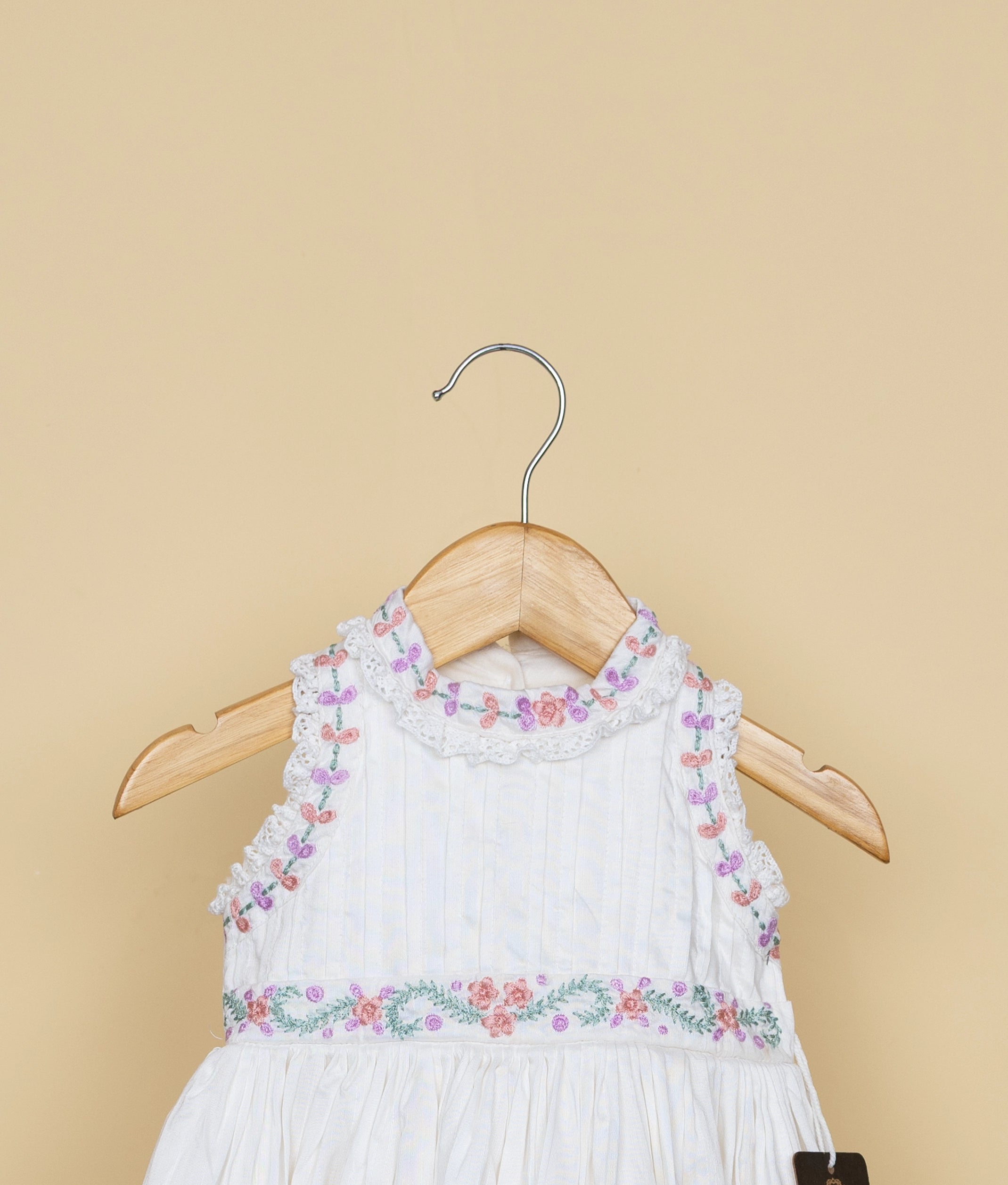 Sleeveless white baby dress with pastel floral embroidery on neck and waist, displayed on wooden hanger against beige background