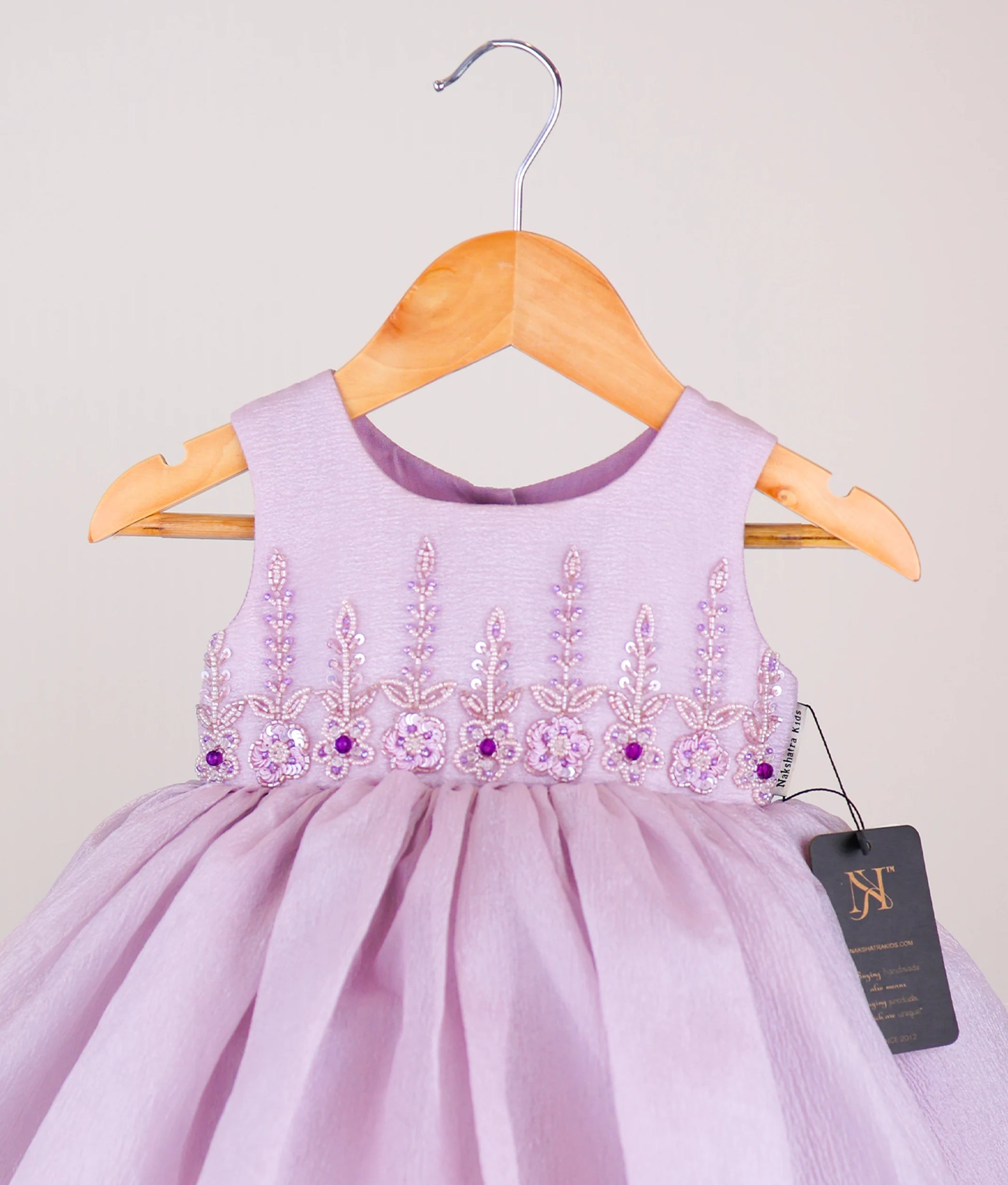 Close view of an pastel lavender frock with floral embroidery on a hanger against a light background.