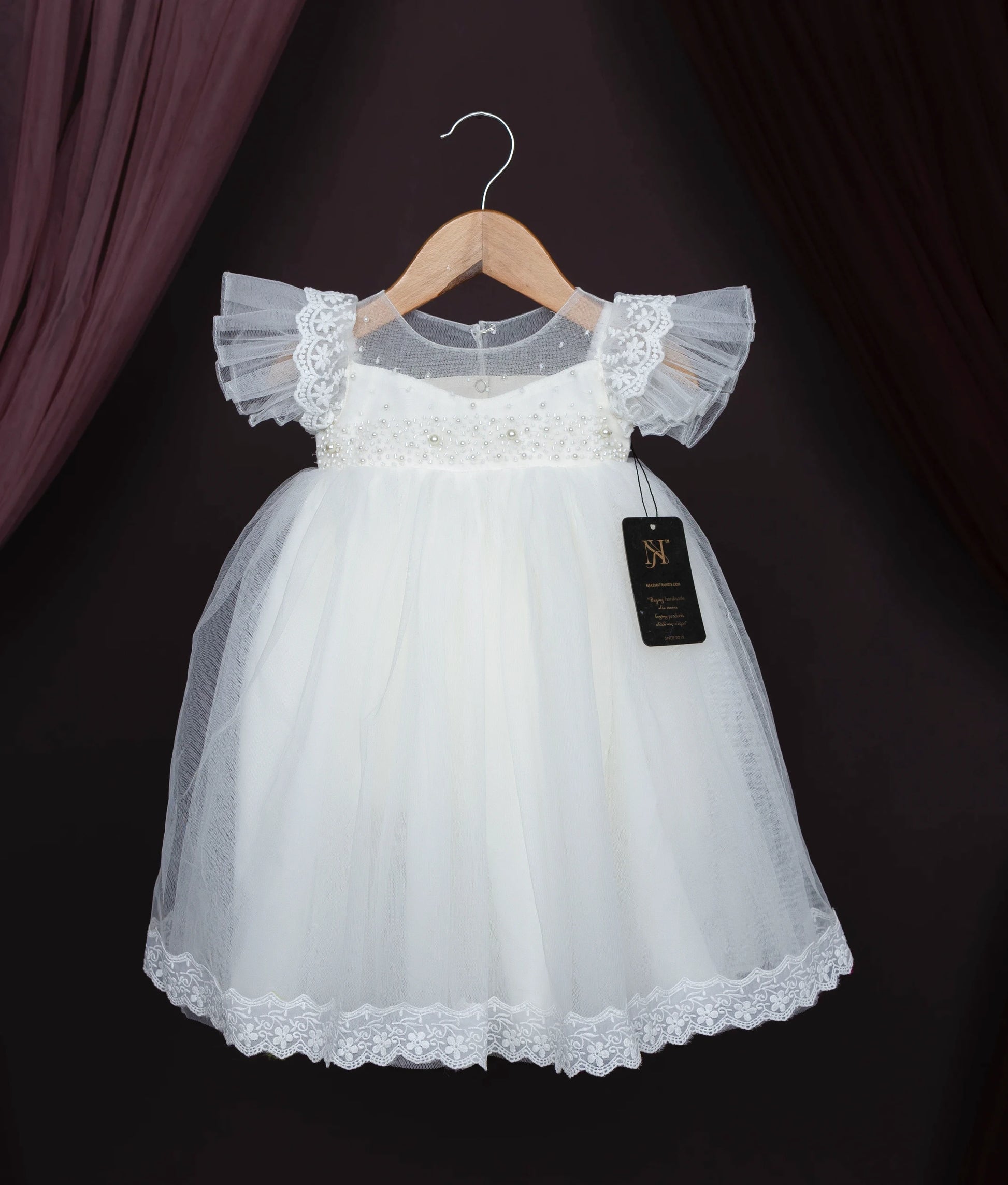Off-white baby girl baptism dress with lace, pearls, and frilled sleeves on a hanger.