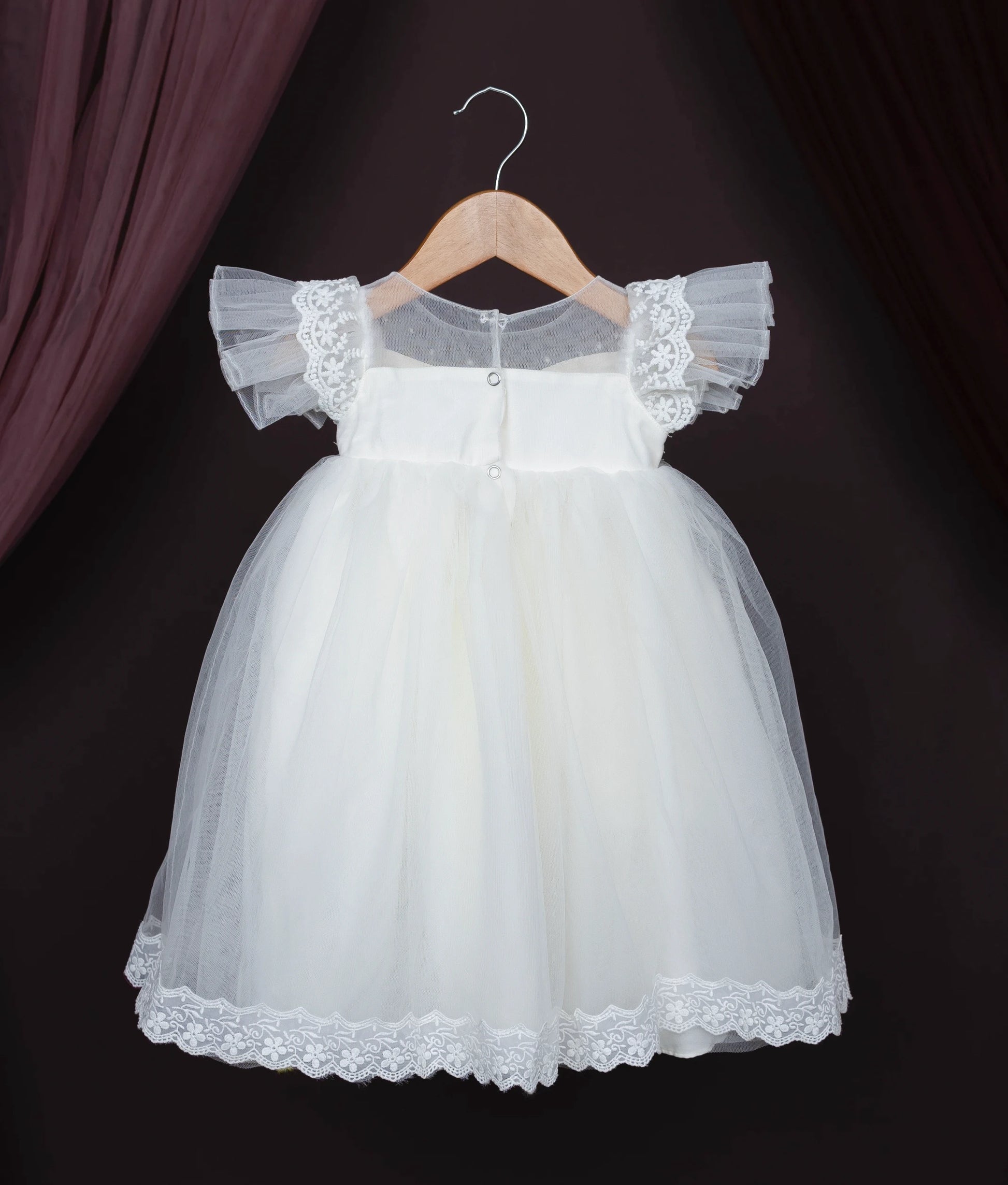 Back view of an off-white baby girl baptism dress with lace, pearls, and frilled sleeves on a hanger.