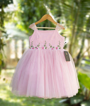 Pastel pink tulle net embroidery baby girl frock hanging on a wooden hanger outdoors.