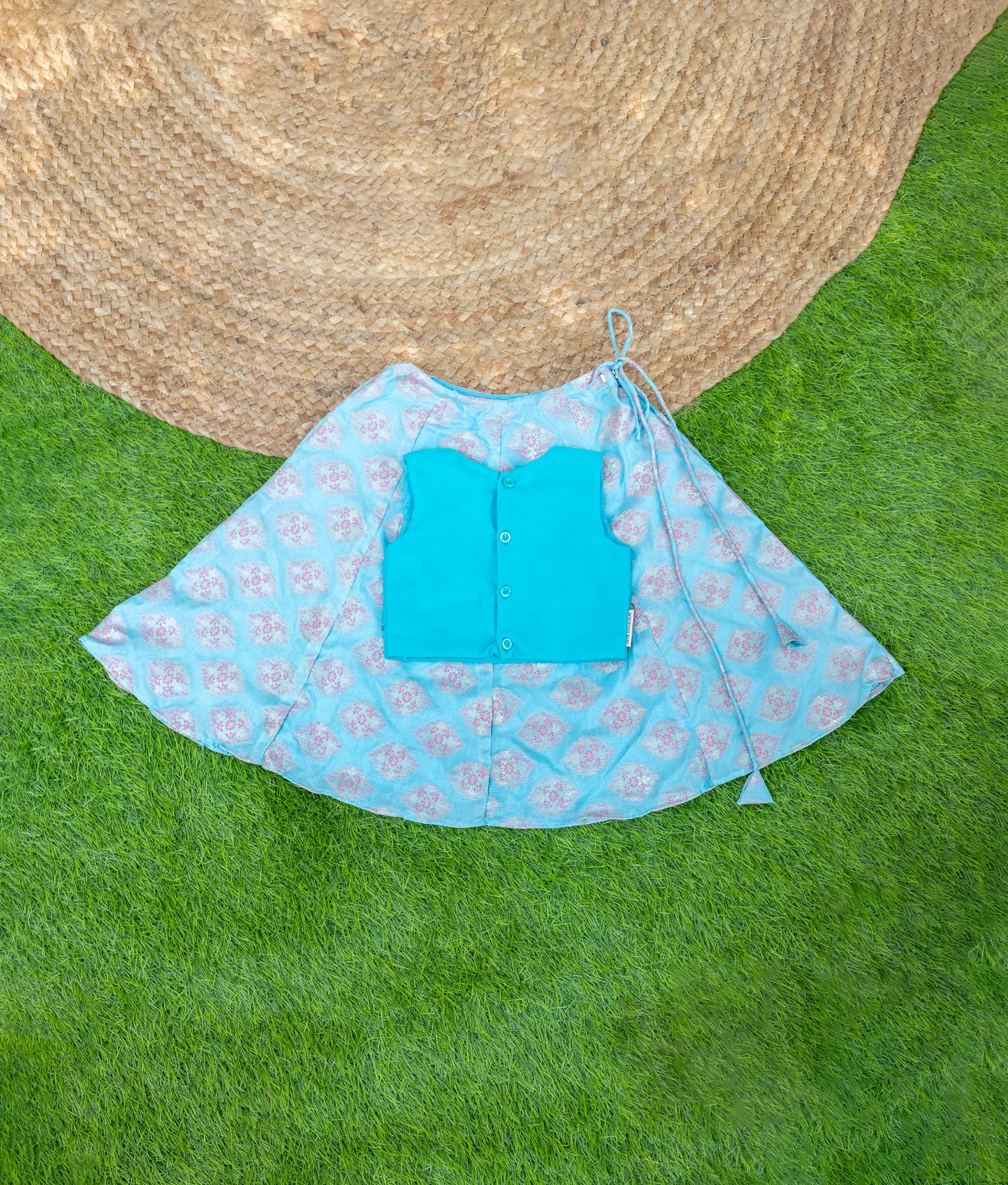 Back view of a traditional outfit displayed on a grass and woven mat background. The outfit features a flared blue skirt with a pink floral pattern and a matching turquoise sleeveless blouse with button details on the back. The skirt has a drawstring tie at the waist.