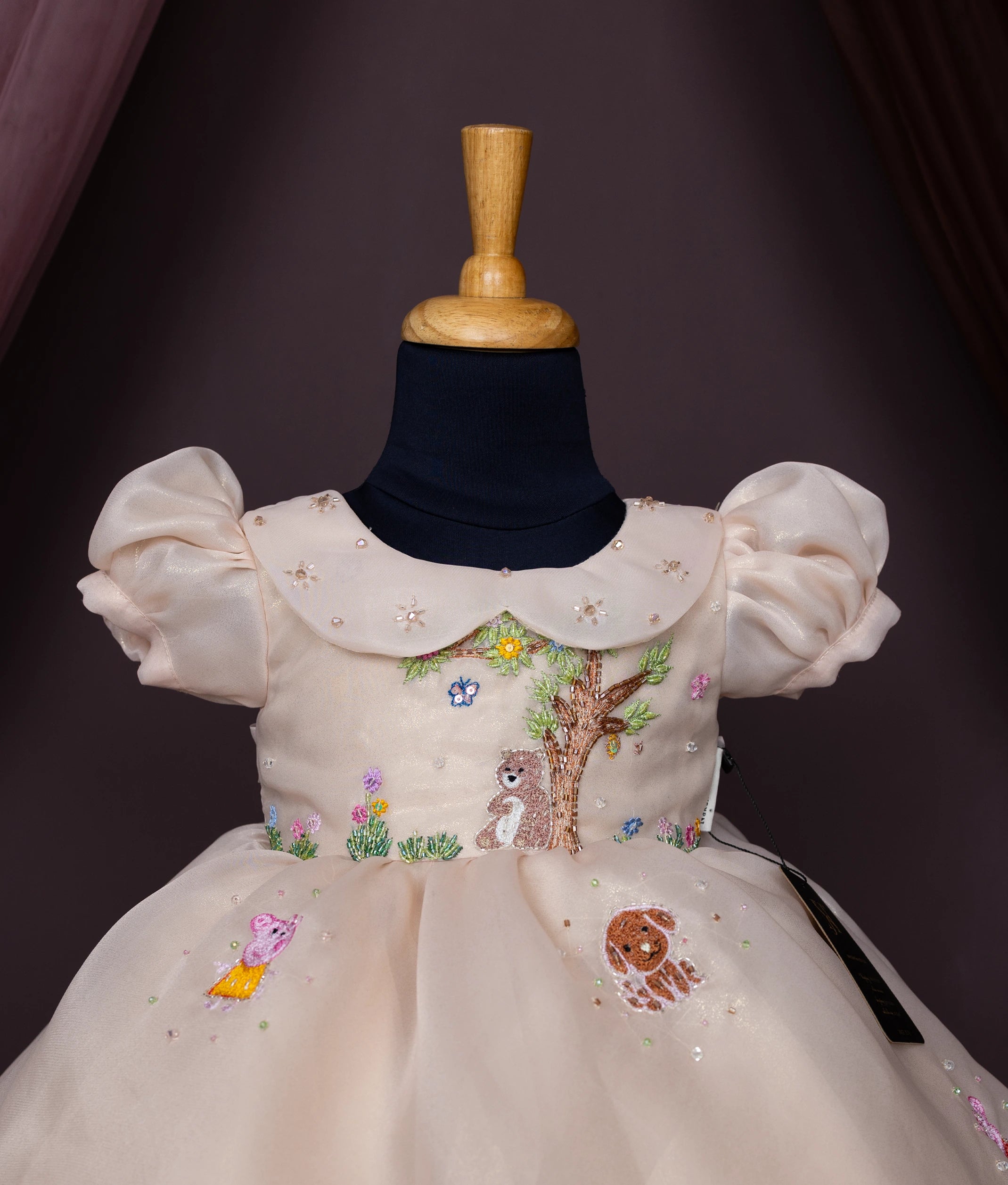 Close view of a beige baby girl dress with puff sleeves, a Peter Pan collar, and cartoon forest animal embroidery.

